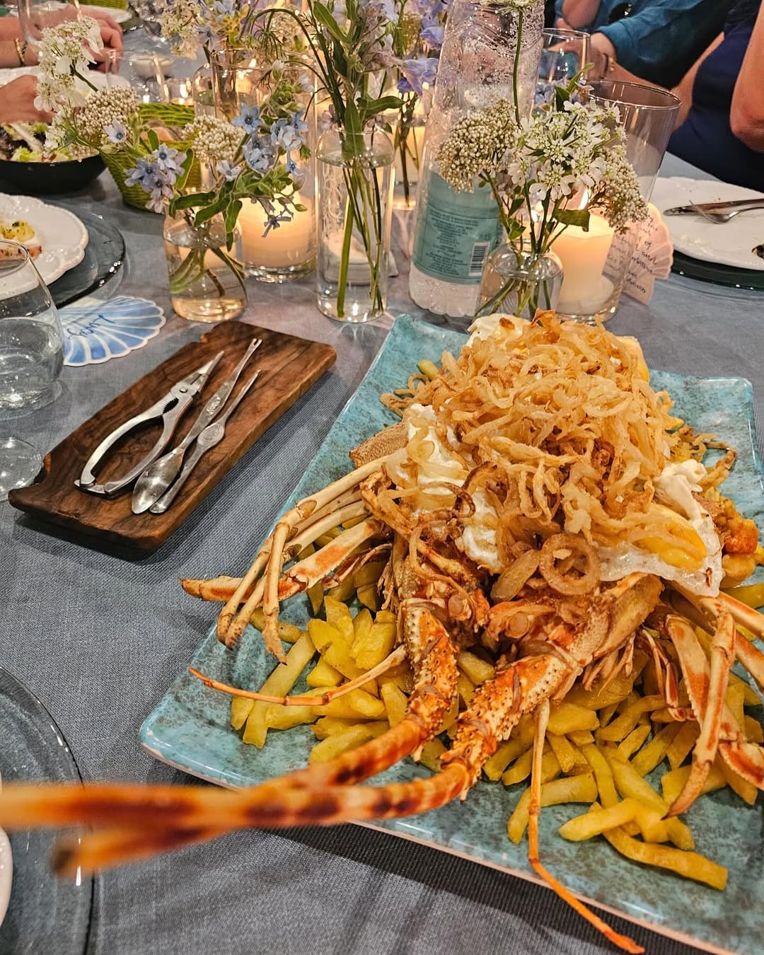 Een hele langoest op friet met gebakken eieren en krokante ui op een turkooizen schaal, met een kreeftentang op een houten plankje ernaast