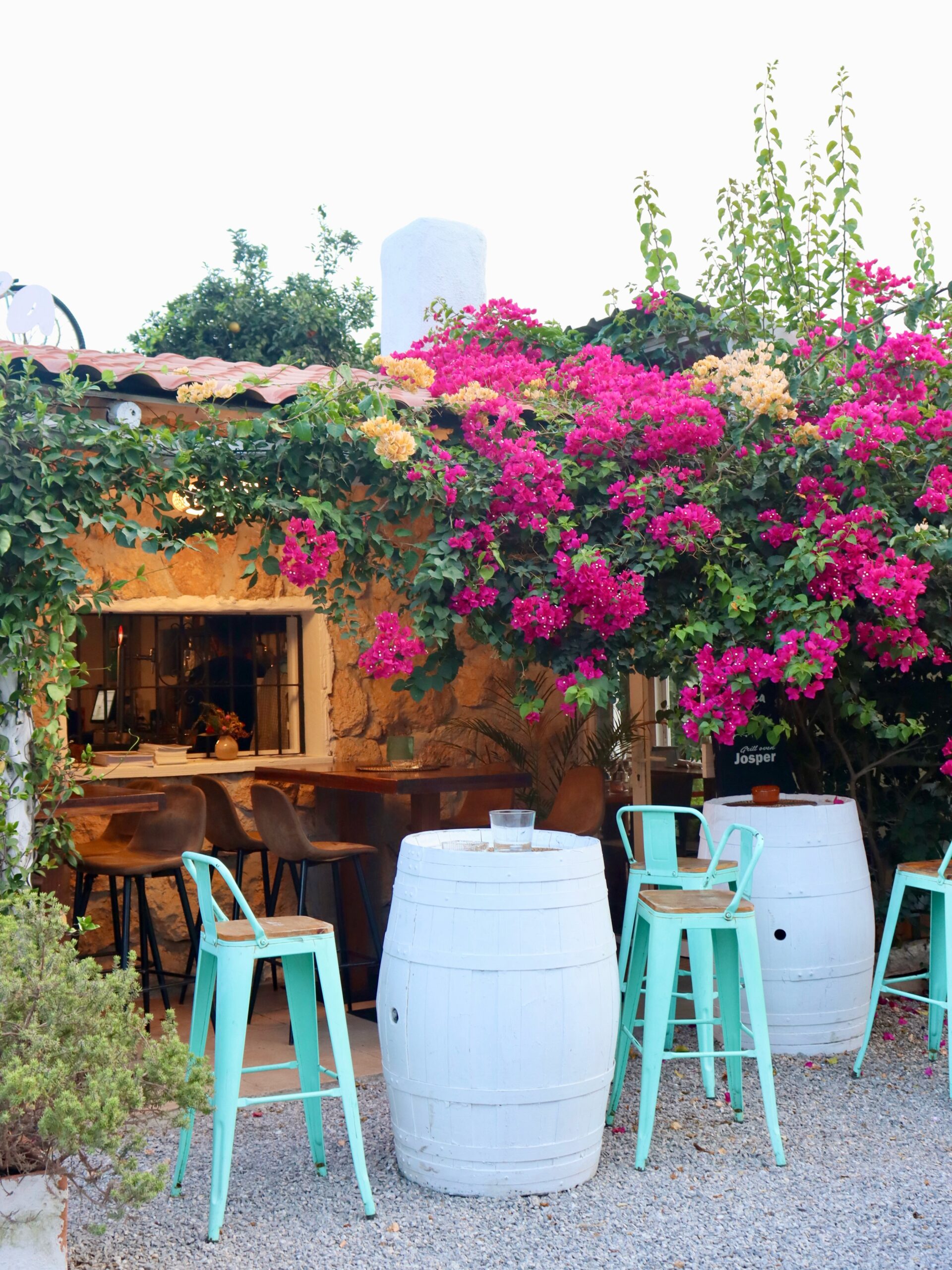 De natuurstenen gevel van Casa Piedra met bougainville over het dak, witte wijnvaten als statafels en een krijtbord met Josper erop
