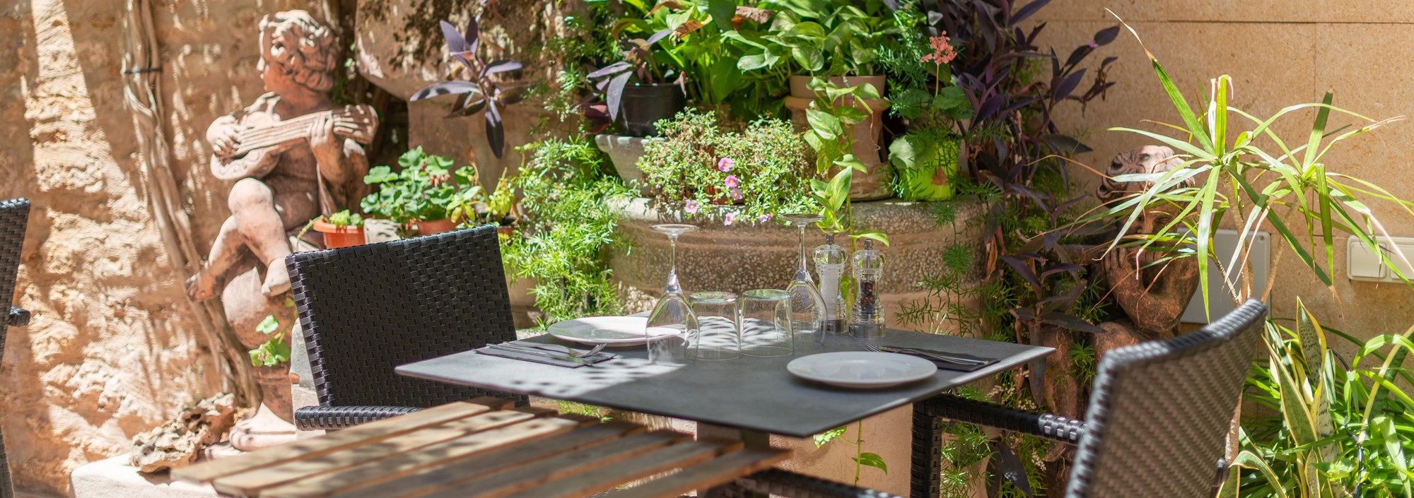 Celler Ca'n Costa in Alcúdia: Mallorquijnse keuken in een huis uit 1594 8 Een gedekte tafel op de binnenplaats van Celler Ca'n Costa in Alcúdia, tegen een natuurstenen muur met een stenen beeld en veel potplanten
