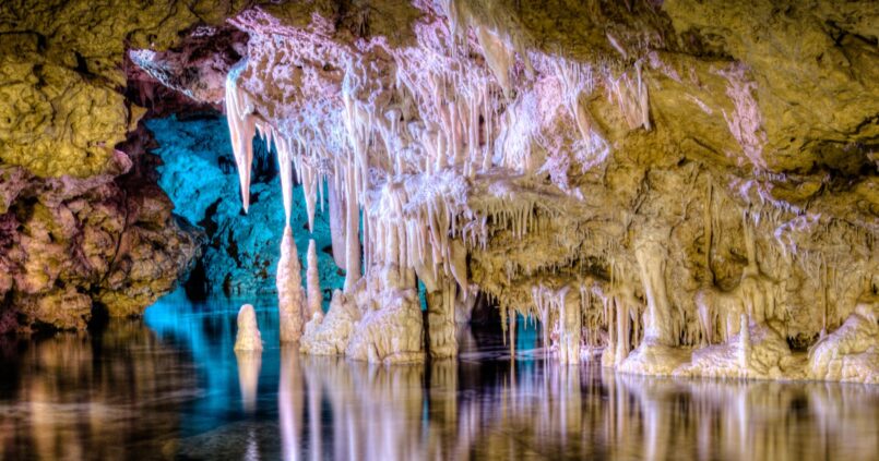Het ondergrondse meer Mar de Venècia in de Coves dels Hams, met blauw en roze uitgelichte druipsteenformaties die in het stille water weerspiegelen