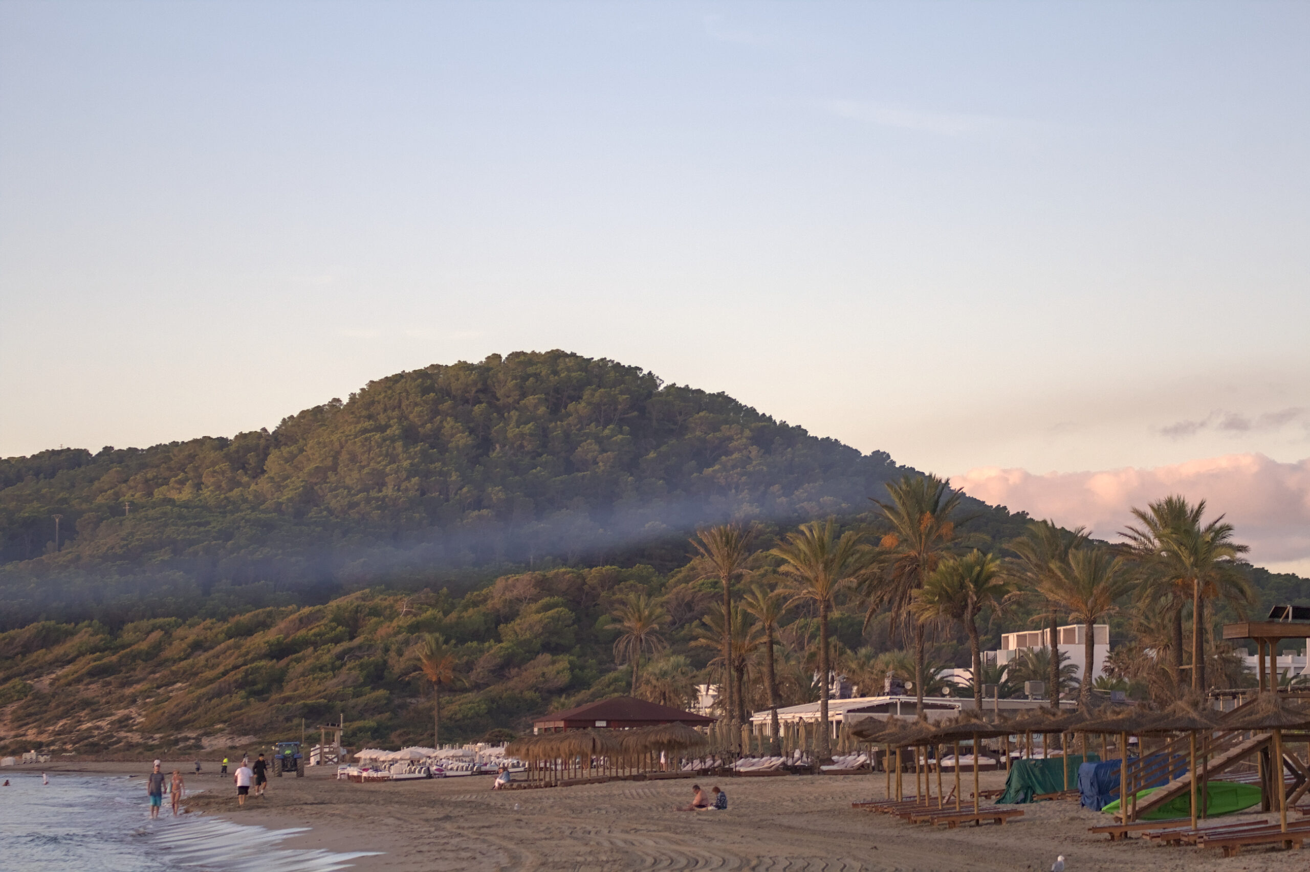 Platja d'en Bossa op Ibiza in het vroege ochtendlicht, met opgestapelde ligbedden en de beboste heuvel erachter