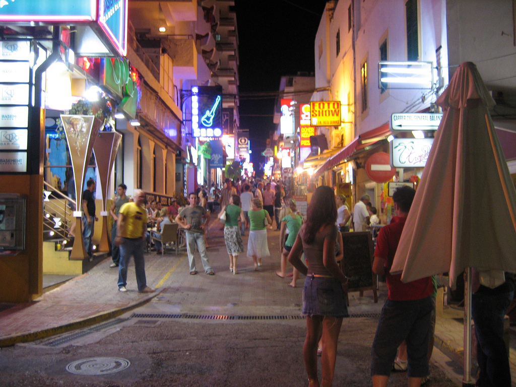 De uitgaansstraat West End in Sant Antoni op Ibiza bij avond, met neonreclames en wandelend publiek
