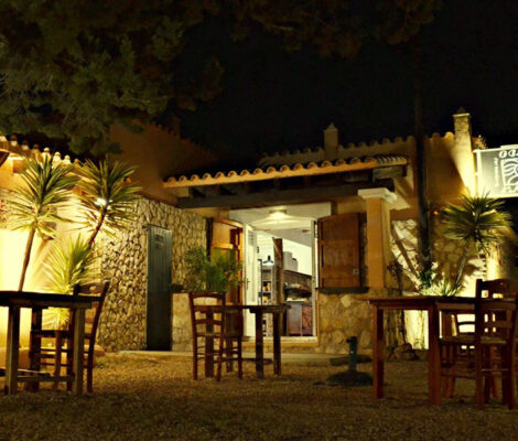 Het verlichte terras van El Cruce by Macondo op Formentera met houten tafels op het grind en palmen