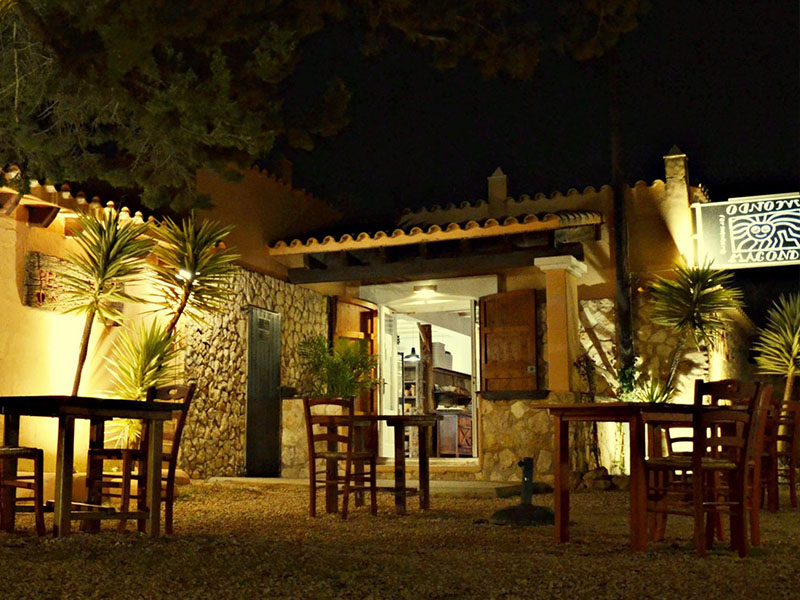 Het verlichte terras van El Cruce by Macondo op Formentera met houten tafels op het grind en palmen