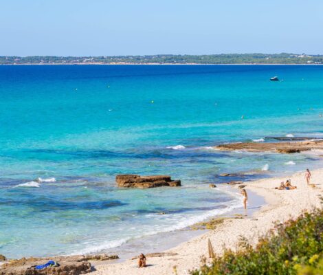 Het brede lichte zand van Es Arenals op Formentera met turquoise water, rotsplaten aan de waterlijn en badgasten op het strand