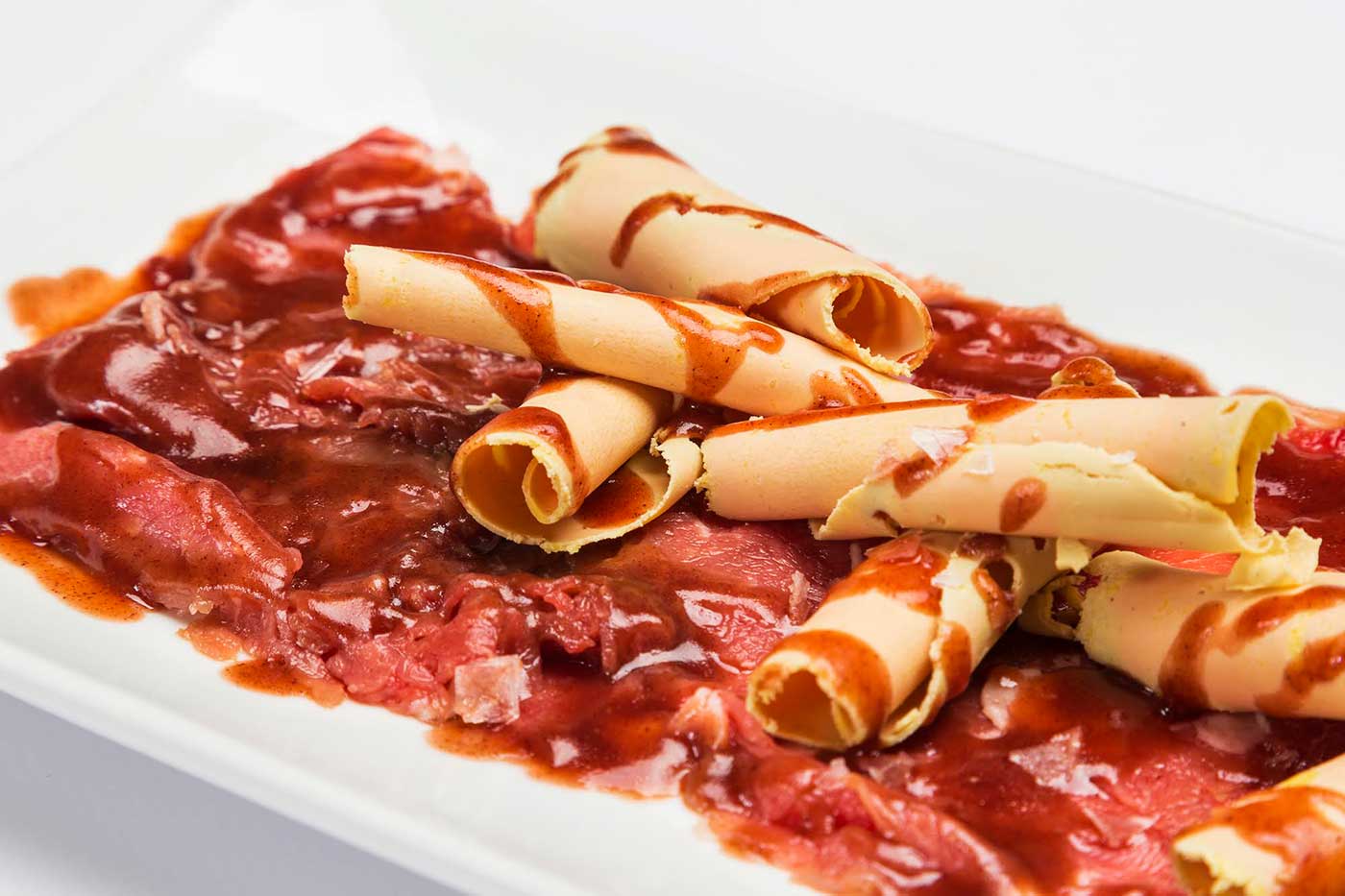 Carpaccio van rundvlees met opgerolde plakjes en een donkere saus op een rechthoekig bord