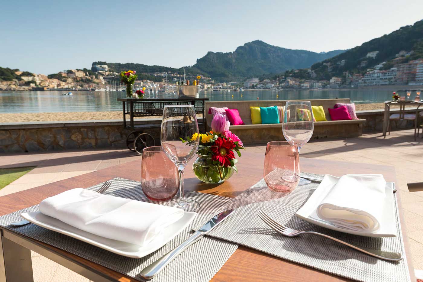 Terrastafel van Es Canyís met bloemen en gekleurde glazen, met uitzicht over de baai en de bergen van de Tramuntana