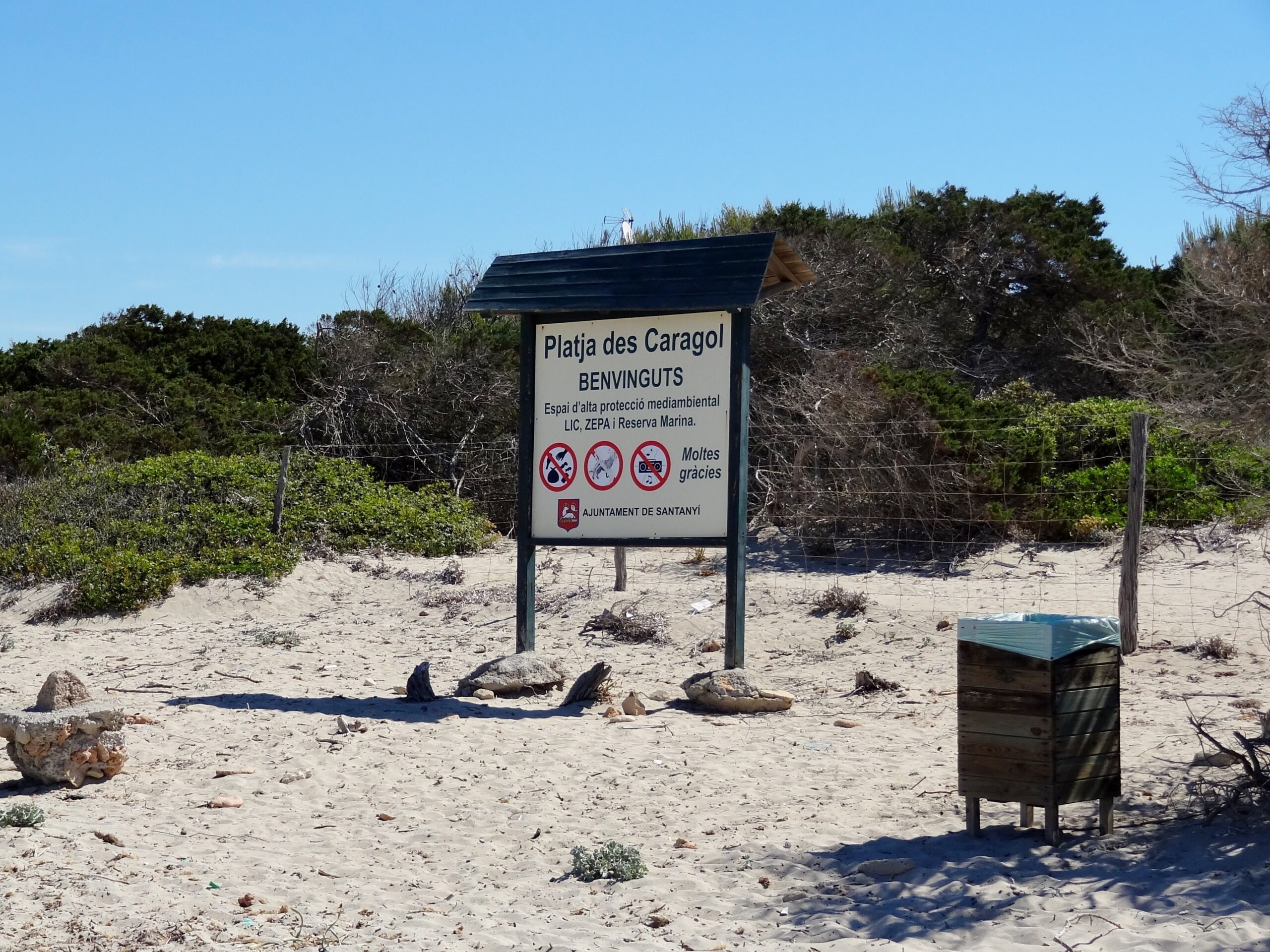 Het welkomstbord bij Es Caragol met de naam Platja des Caragol en de regels voor dit beschermde natuurgebied, tussen de duinstruiken op het zand