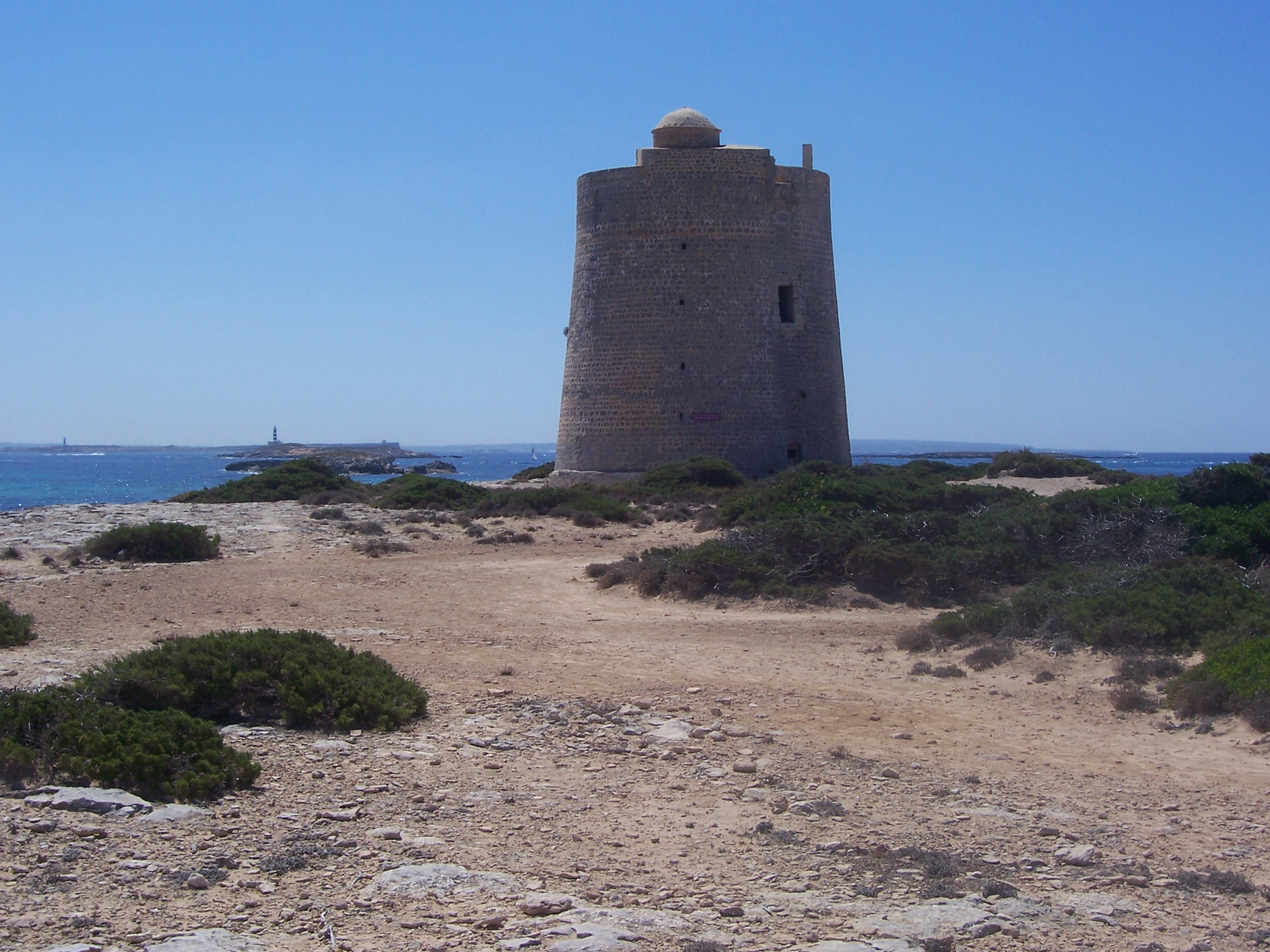 Het zandpad over de landtong aan het zuideinde van Es Cavallet naar de ronde wachttoren Torre de ses Portes, met lage struiken en op de achtergrond de eilandjes in Es Freus