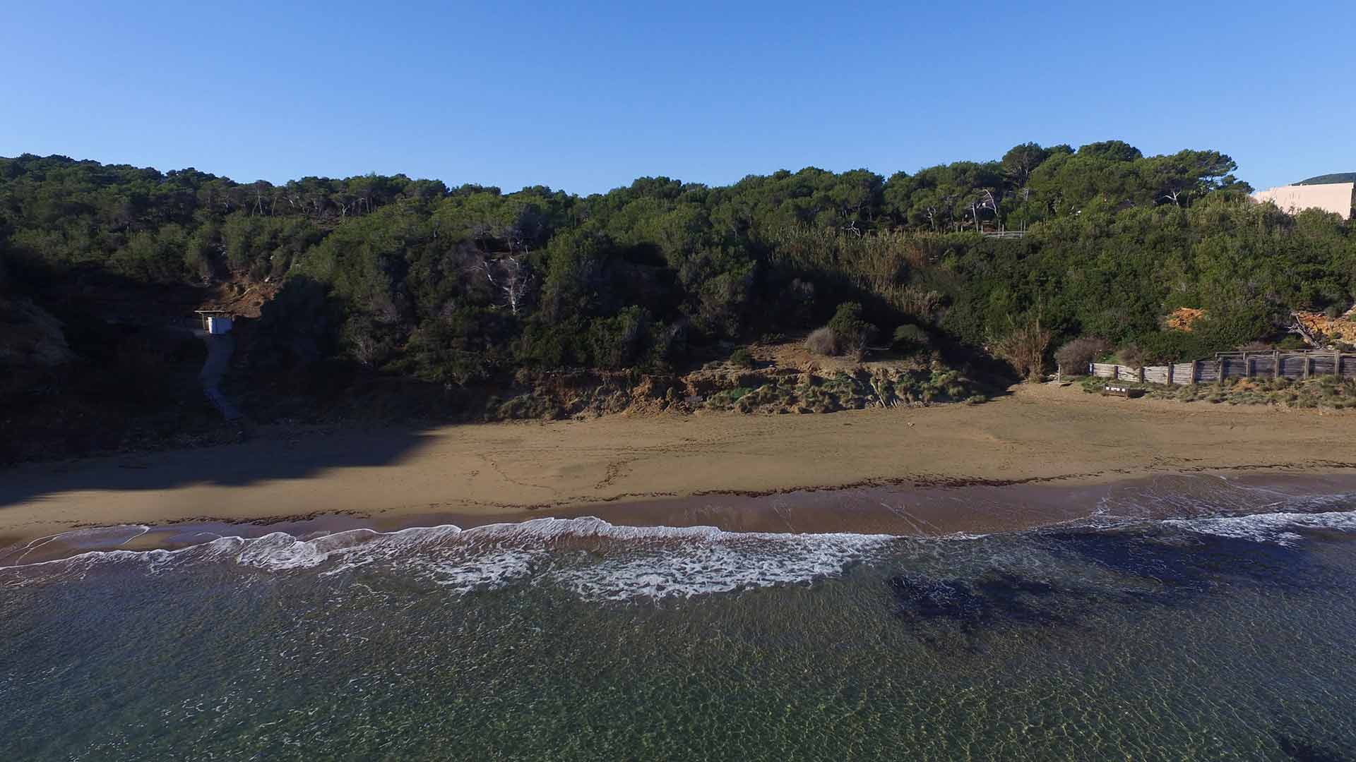 Es Figueral buiten het seizoen: een leeg zandstrand met branding, de beboste helling erachter en rechts de houten afrastering langs de loopplanken