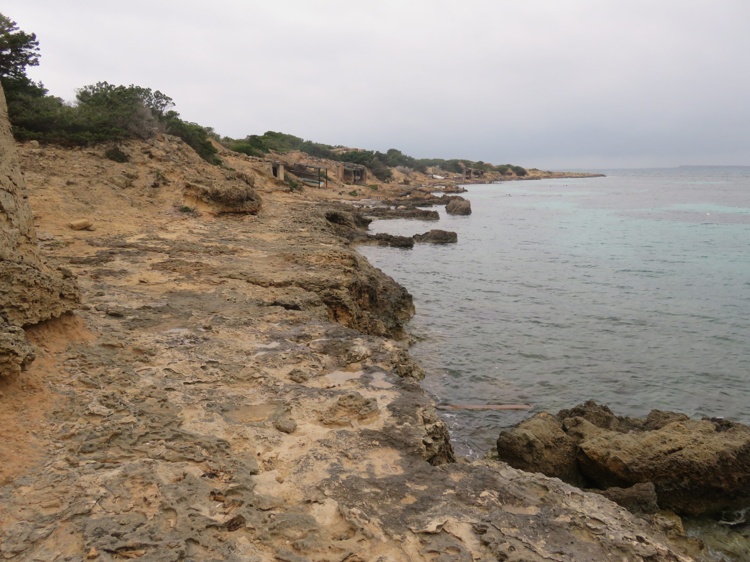 De rotsige kust bij Caló de s'Oli op Formentera onder een grijze aprilhemel, met in de verte de houten bootgarages