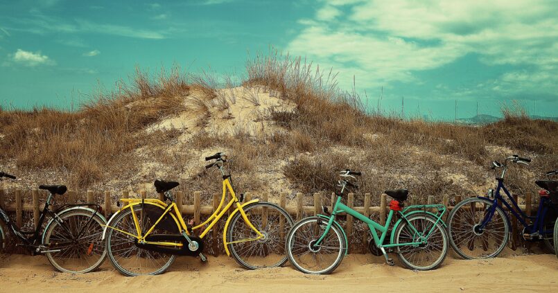 Vier gekleurde fietsen tegen een houten duinhek op Formentera in juli, met droog duingras en een blauwe lucht erachter