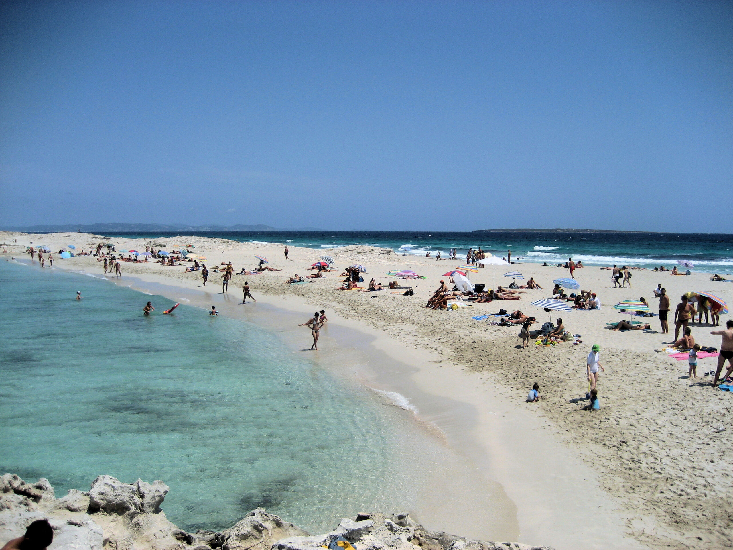 De zandtong van Es Trucadors op Formentera in juli, met badgasten op het zand en helder turkoois water aan weerskanten