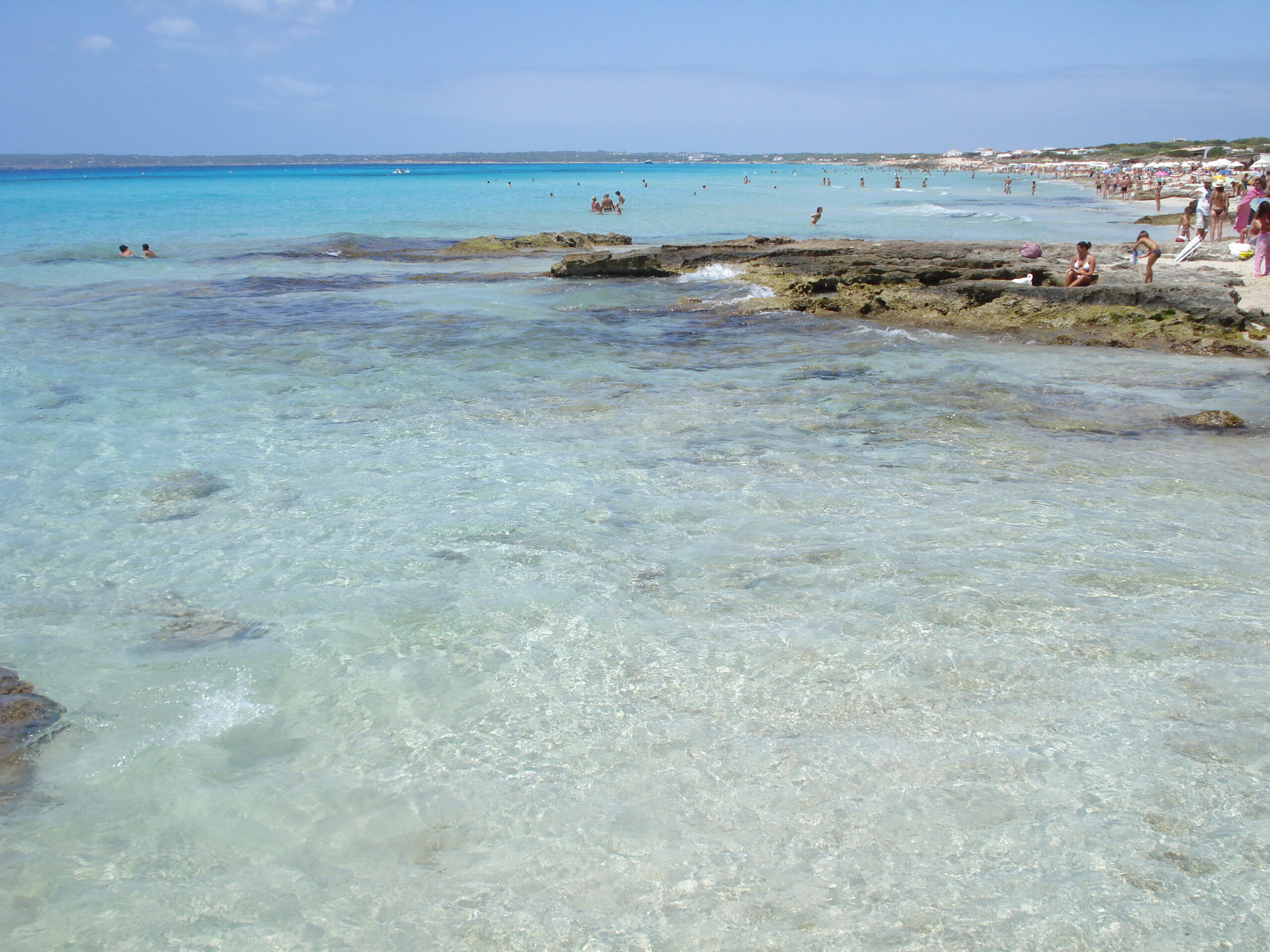 Formentera in juni: zeewater, lange dagen en het voertuigquotum 2 Helder ondiep water over vlakke rotsen bij Es Copinar aan Platja de Migjorn op Formentera met badgasten op de achtergrond