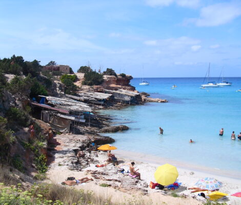 Cala Saona op Formentera in mei, met de houten bootgarages, een paar parasols op het zand en zeilboten voor de kust