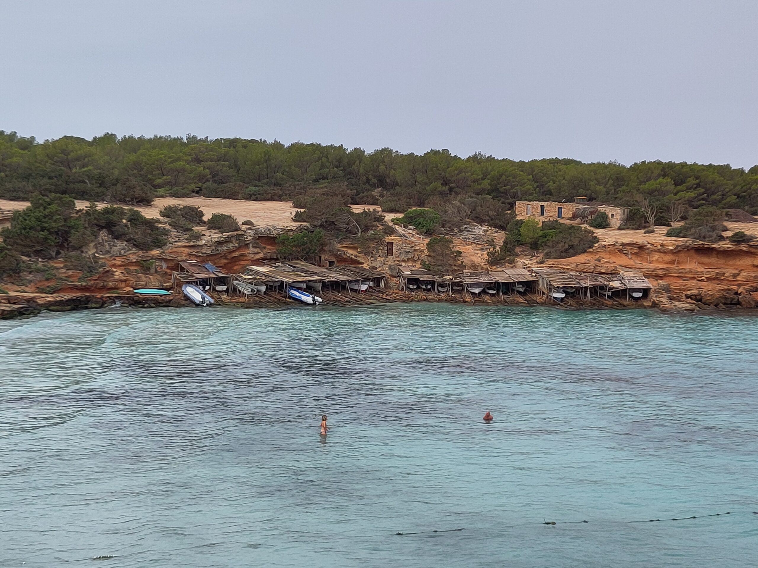 Formentera in september: het warmste zeewater en een leger eiland 2 De oude bootgarages van Cala Saona op Formentera onder een grijze lucht met een enkele zwemmer in het water
