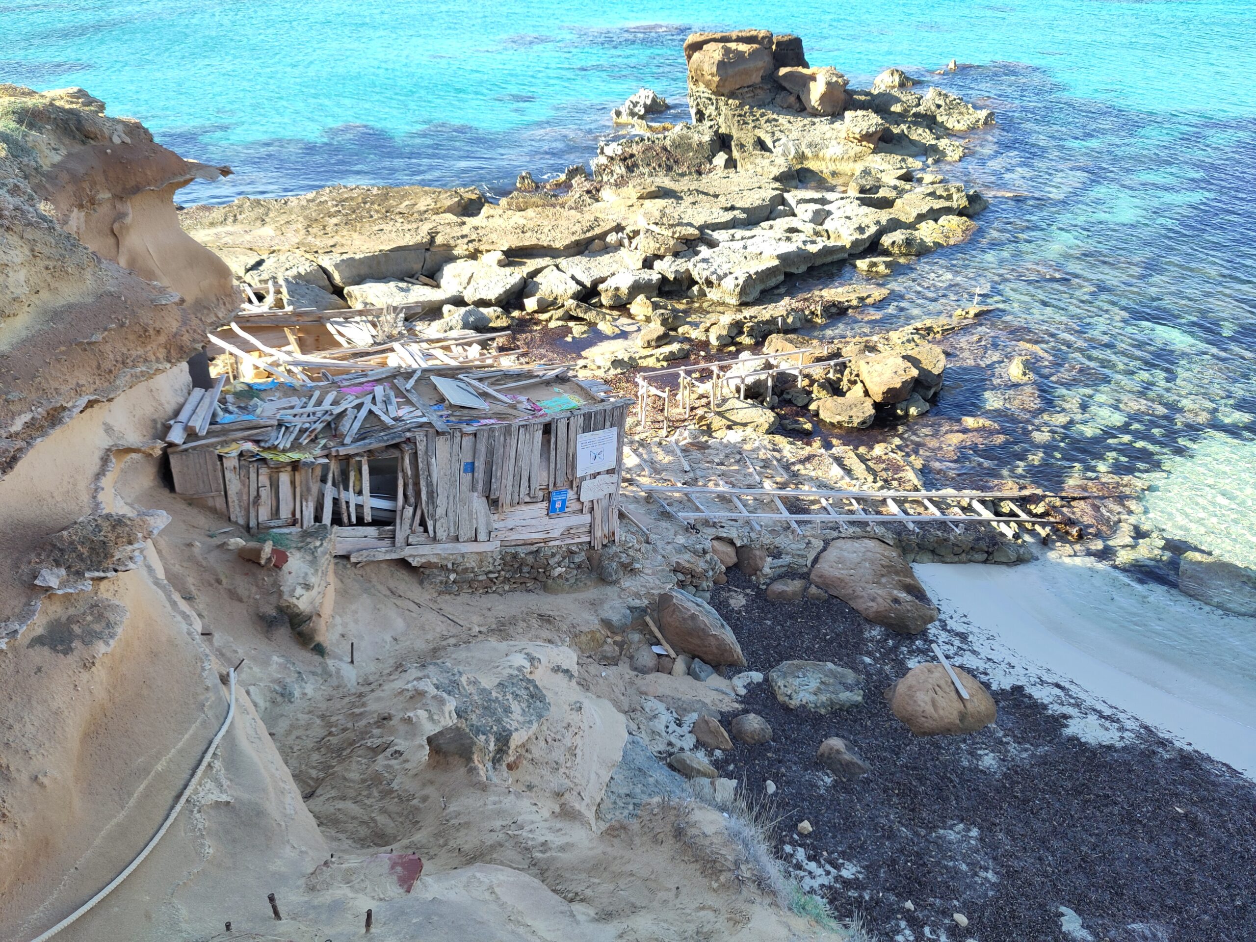 De eenzame houten boothelling van Es Calo des Mort tussen de zandsteenrotsen, met een smalle strook zand en glashelder turquoise water