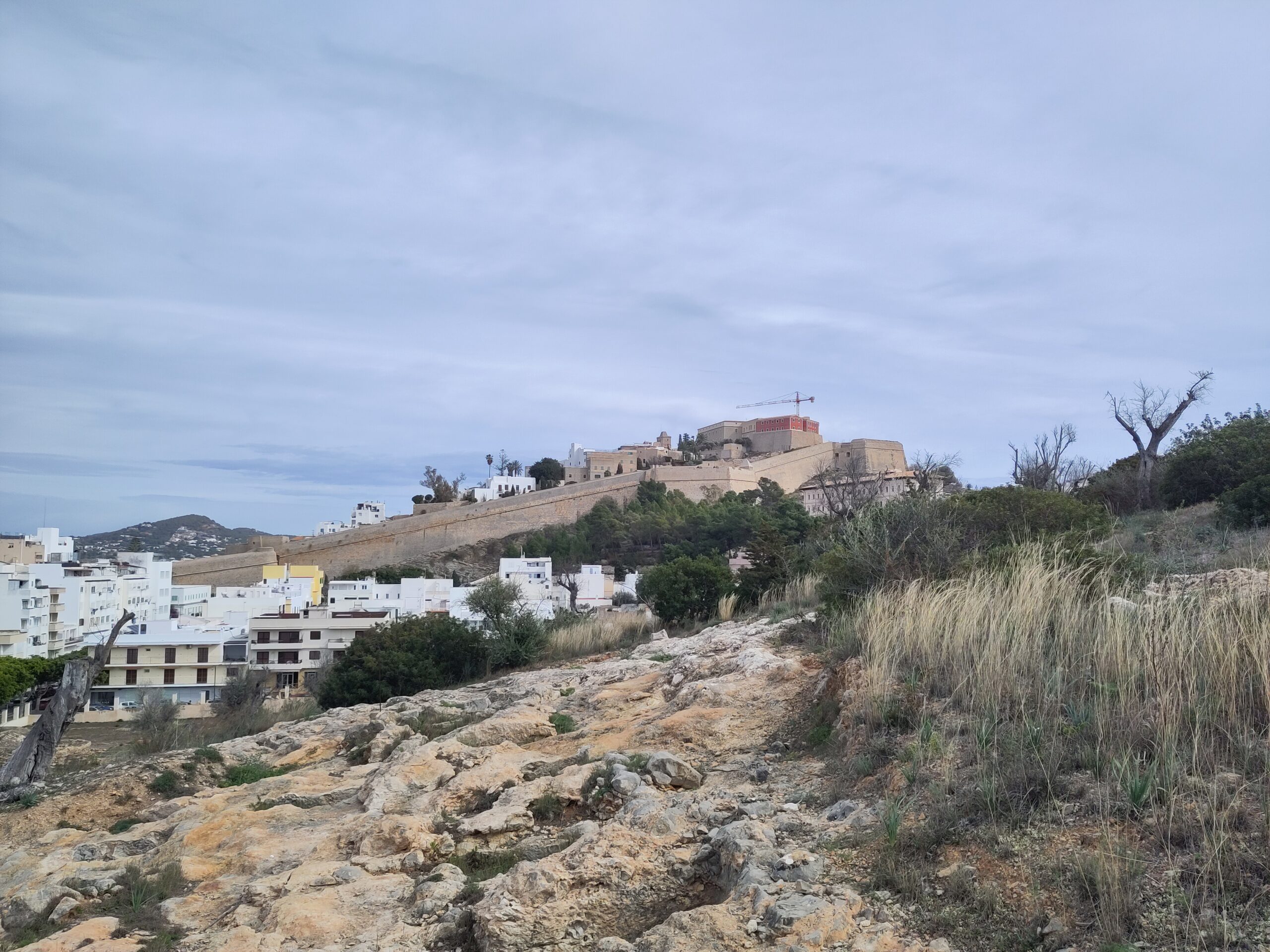 Ibiza in de winter: weer, zeewater en wat er doorgaat 1 De vestingmuren van Dalt Vila in Ibiza-stad onder een bewolkte februarihemel, met droog gras op de voorgrond