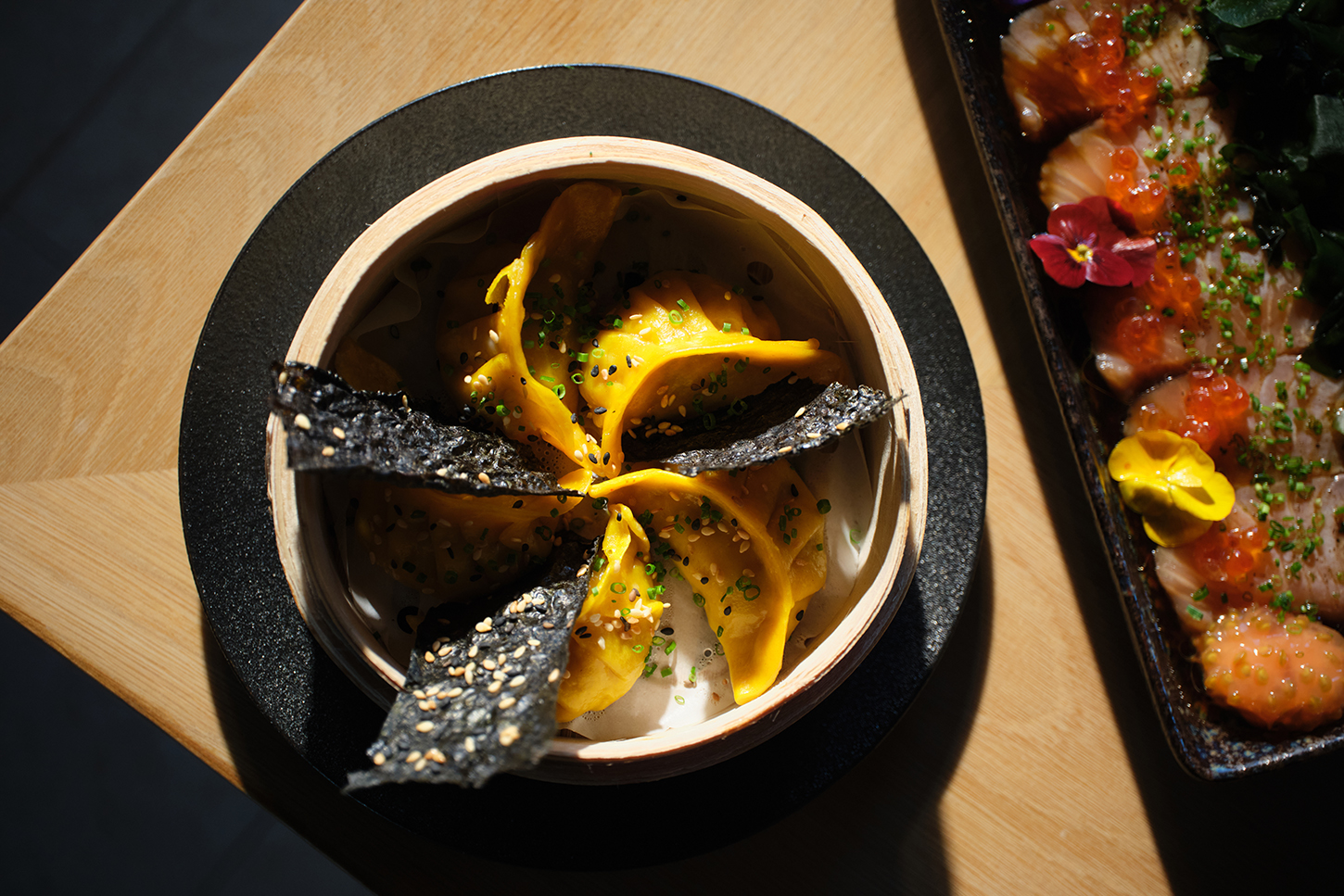 Gele dumplings in een bamboestomer met geroosterd zeewier, naast een schaal sashimi met viskuit en een eetbare bloem bij KATAGI Blau