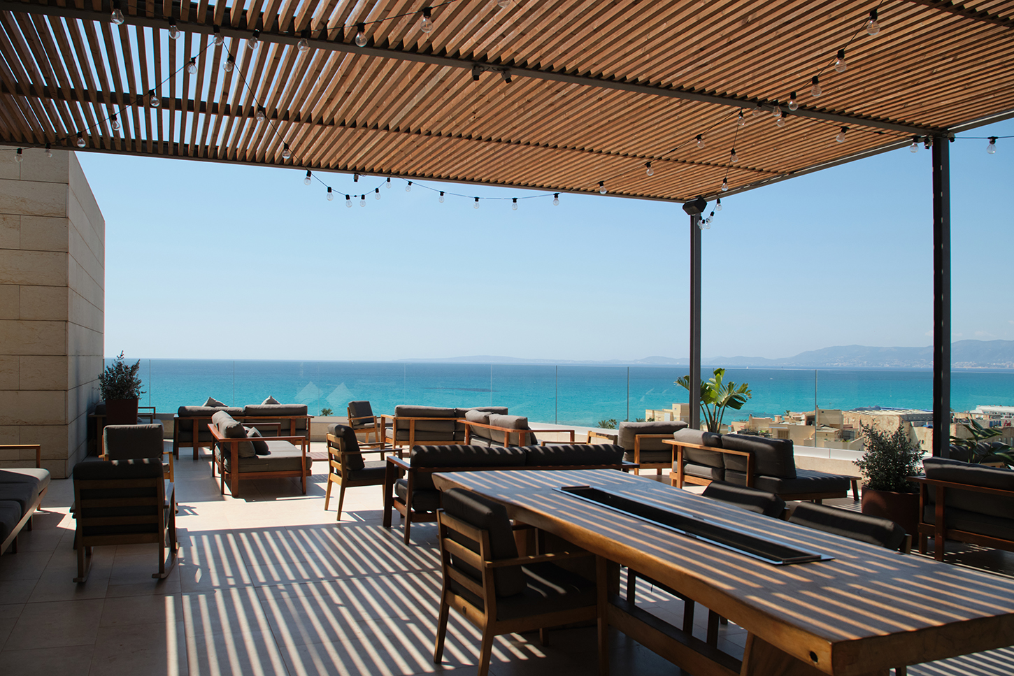 Loungehoek van het dakterras van KATAGI Blau overdag, met houten pergola, lichtsnoeren en banken met uitzicht op het strand van Playa de Palma