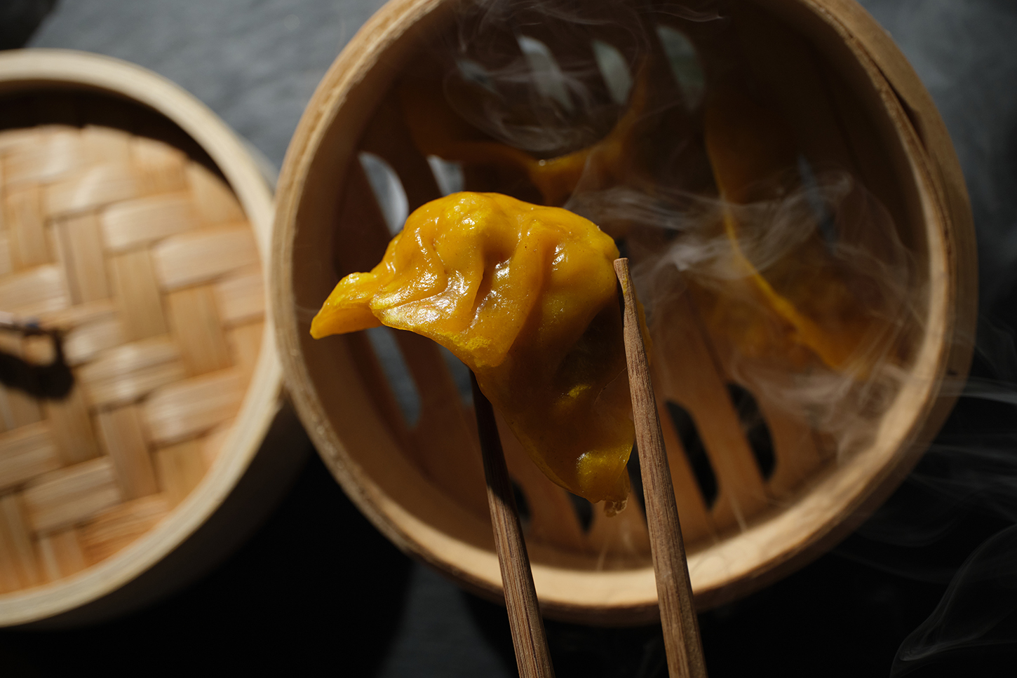 Een gele dumpling tussen stokjes boven een dampende bamboestomer bij KATAGI Blau