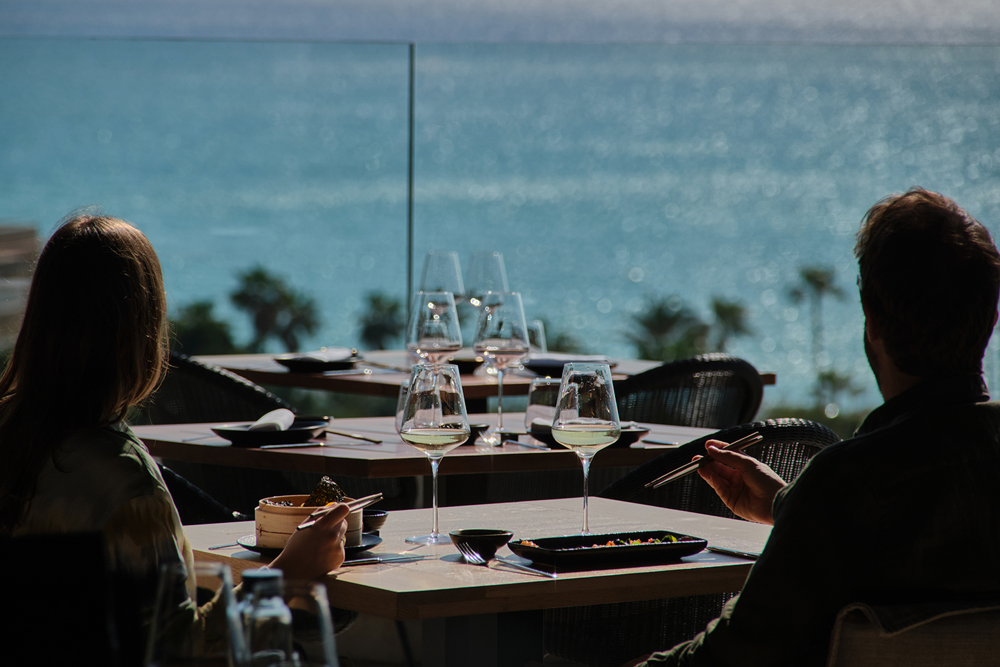 Twee gasten van achteren gezien aan een tafel op het terras van KATAGI Blau, met wijnglazen en kleine schalen en de zee op de achtergrond