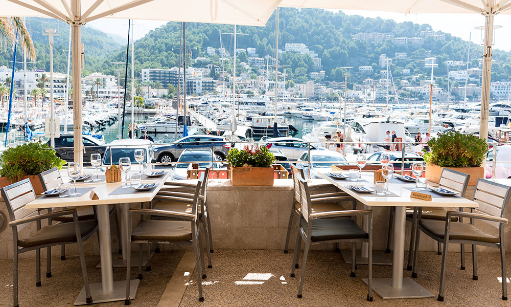 Kingfisher Port de Sóller: vis en zeevruchten boven de jachthaven 6 Het terras van Kingfisher bij daglicht, met witte parasols, gedekte tafels en daarachter de steigers van de jachthaven van Port de Sóller