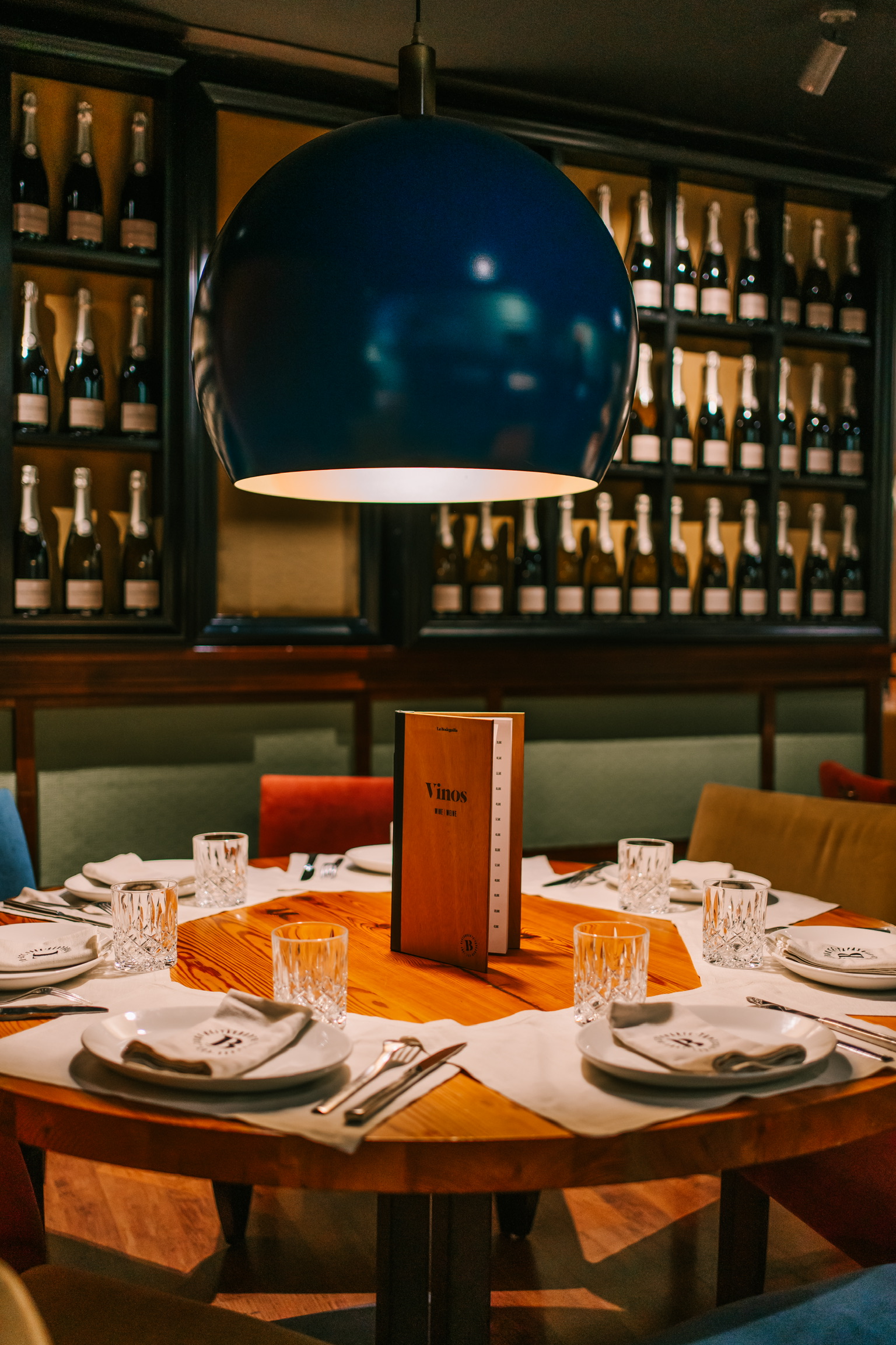 Gedekte ronde tafel bij La Bodeguilla in Palma onder een blauwe hanglamp, met de wijnwand op de achtergrond