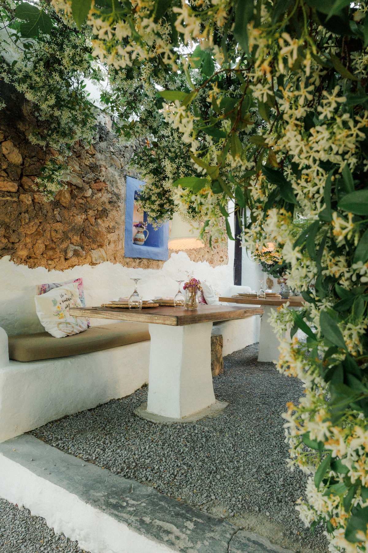 La Luna nell’Orto Ibiza: zelfgemaakte pasta onder een eeuwenoude vijgenboom 4 Gemetselde witte bank met kussens en een houten tafel onder bloeiende jasmijn tegen een ruwe stenen muur bij La Luna nell’Orto