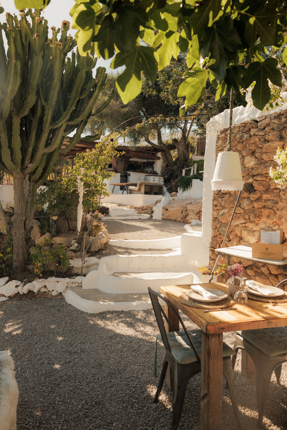 La Luna nell’Orto Ibiza: zelfgemaakte pasta onder een eeuwenoude vijgenboom 2 Tuin van La Luna nell’Orto met witgekalkte trappen tussen de niveaus, een droge stenen muur, cactussen en een gedekte tafel op het grind
