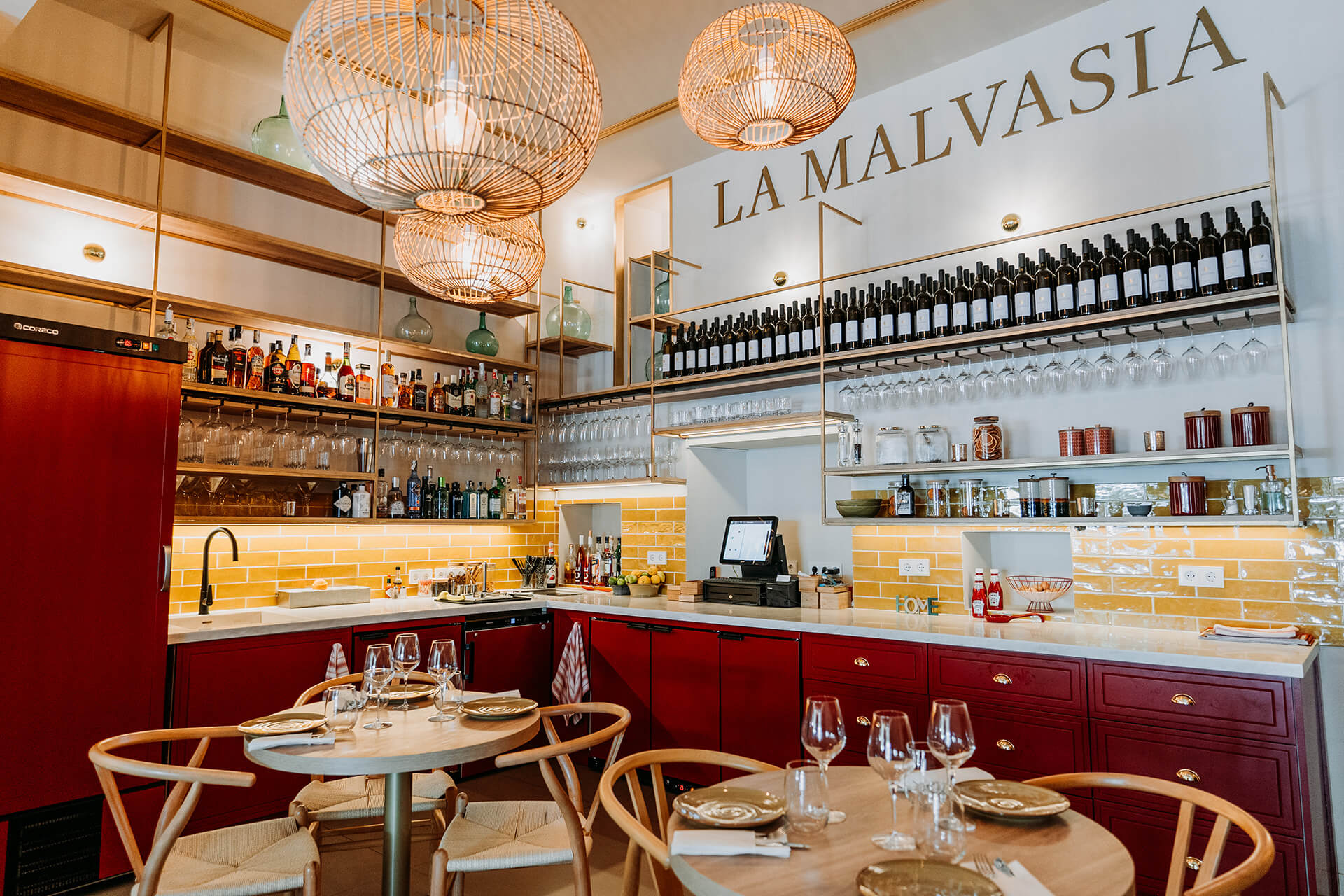 De binnenruimte van restaurant La Malvasia in Palma met gele tegels, bordeauxrode kasten, rieten lampen en de naam in grote letters boven de bar