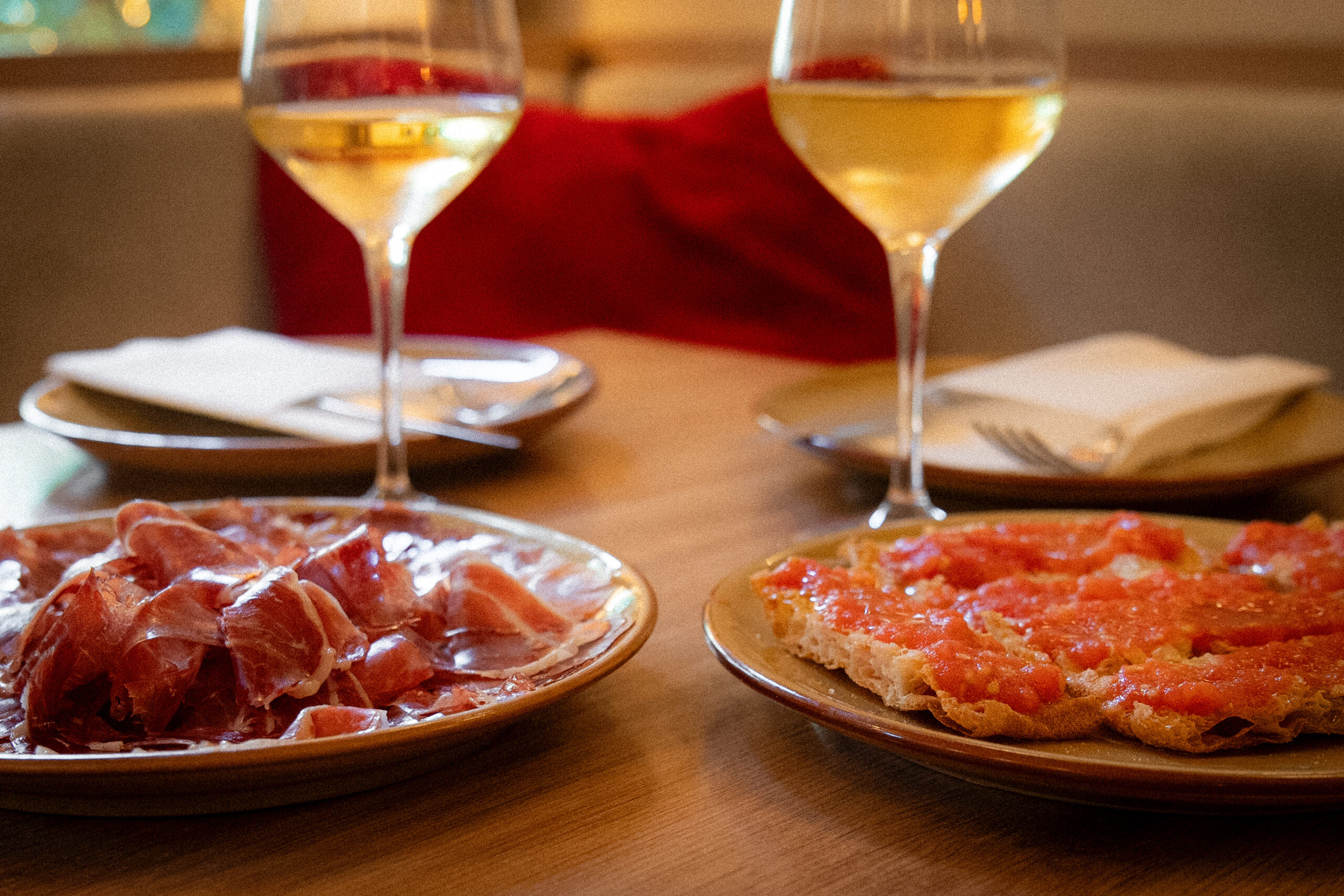 Een bord ibericoham en een geroosterde snee brood met tomaat naast twee glazen witte wijn op een houten tafel bij La Malvasia in Palma