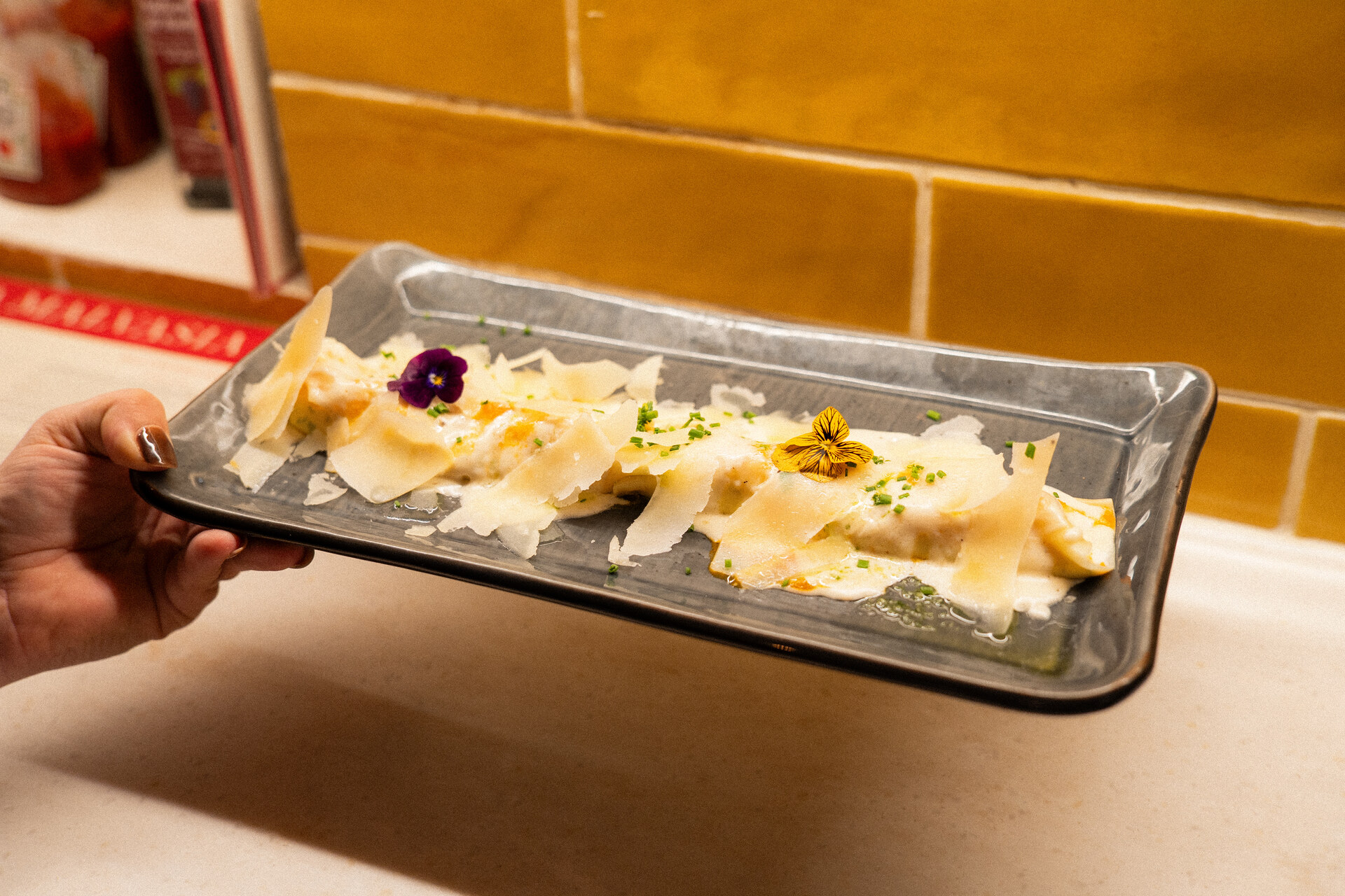 Een langwerpig bord ravioli met schilfers parmezaanse kaas en eetbare bloemen, gedragen voor een gele tegelwand