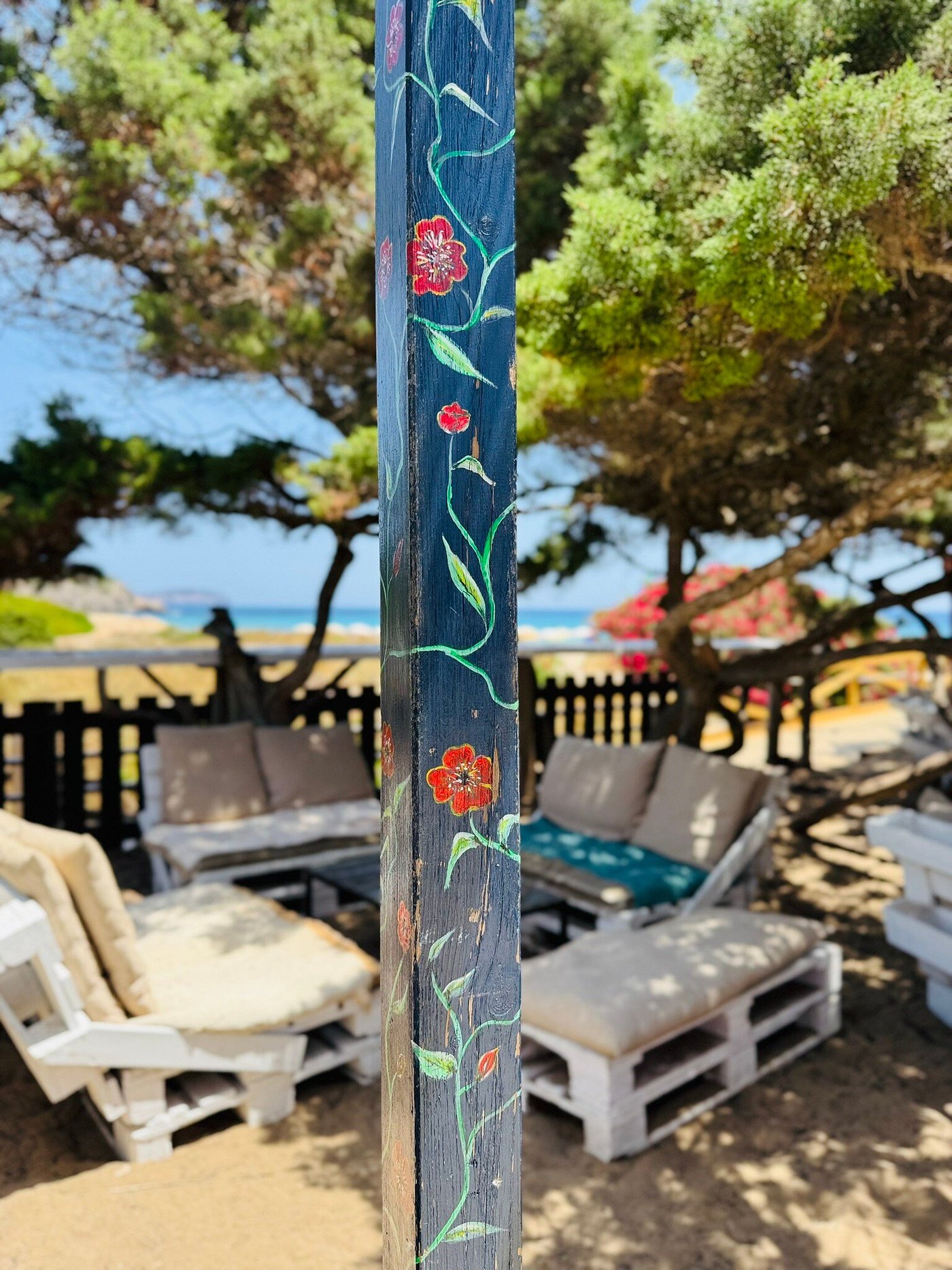 L'Amorigen Ibiza: het beschilderde strandrestaurant van Cala Nova 2 Een met de hand beschilderde donkerblauwe paal met klaprozen en ranken bij L'Amorigen, met daarachter banken van pallets met kussens, pijnbomen en de zee