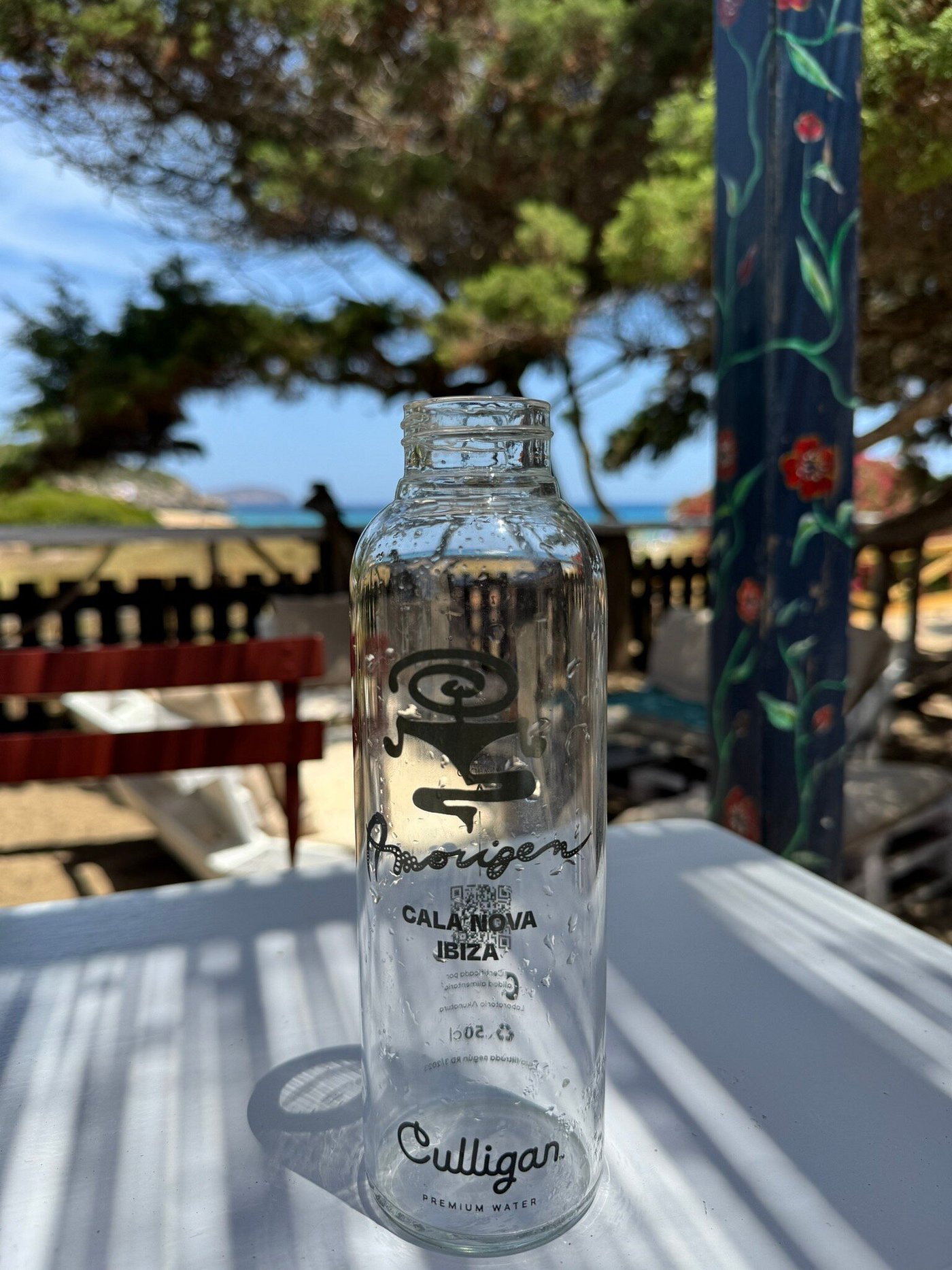 L'Amorigen Ibiza: het beschilderde strandrestaurant van Cala Nova 7 Een glazen waterfles met het etiket van L'Amorigen Cala Nova op tafel, met de beschilderde paal en de baai op de achtergrond
