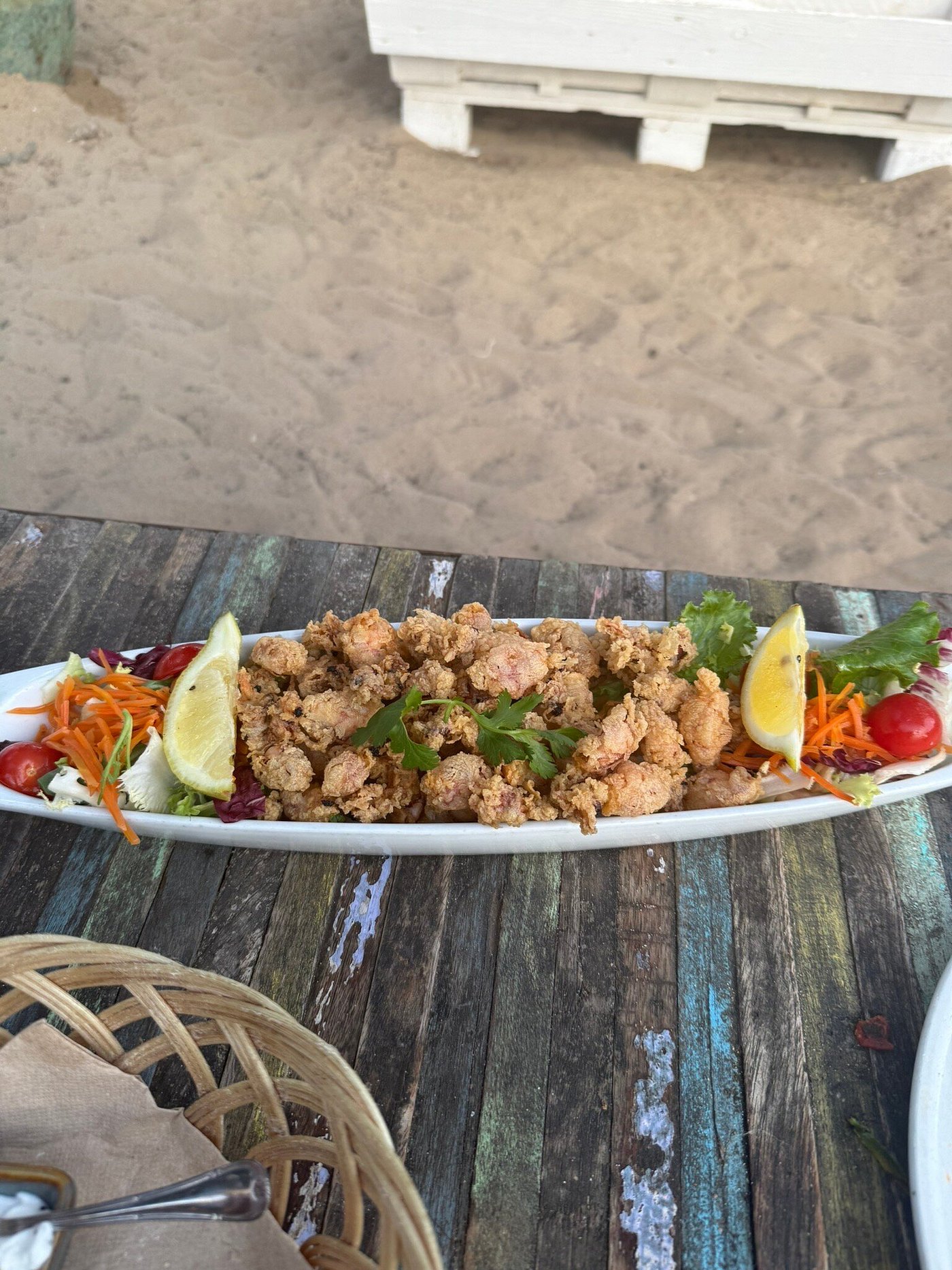 L'Amorigen Ibiza: het beschilderde strandrestaurant van Cala Nova 5 Een langwerpig bord met gefrituurde inktvis en citroen op een verweerde houten tafel, met het zand van Cala Nova erachter