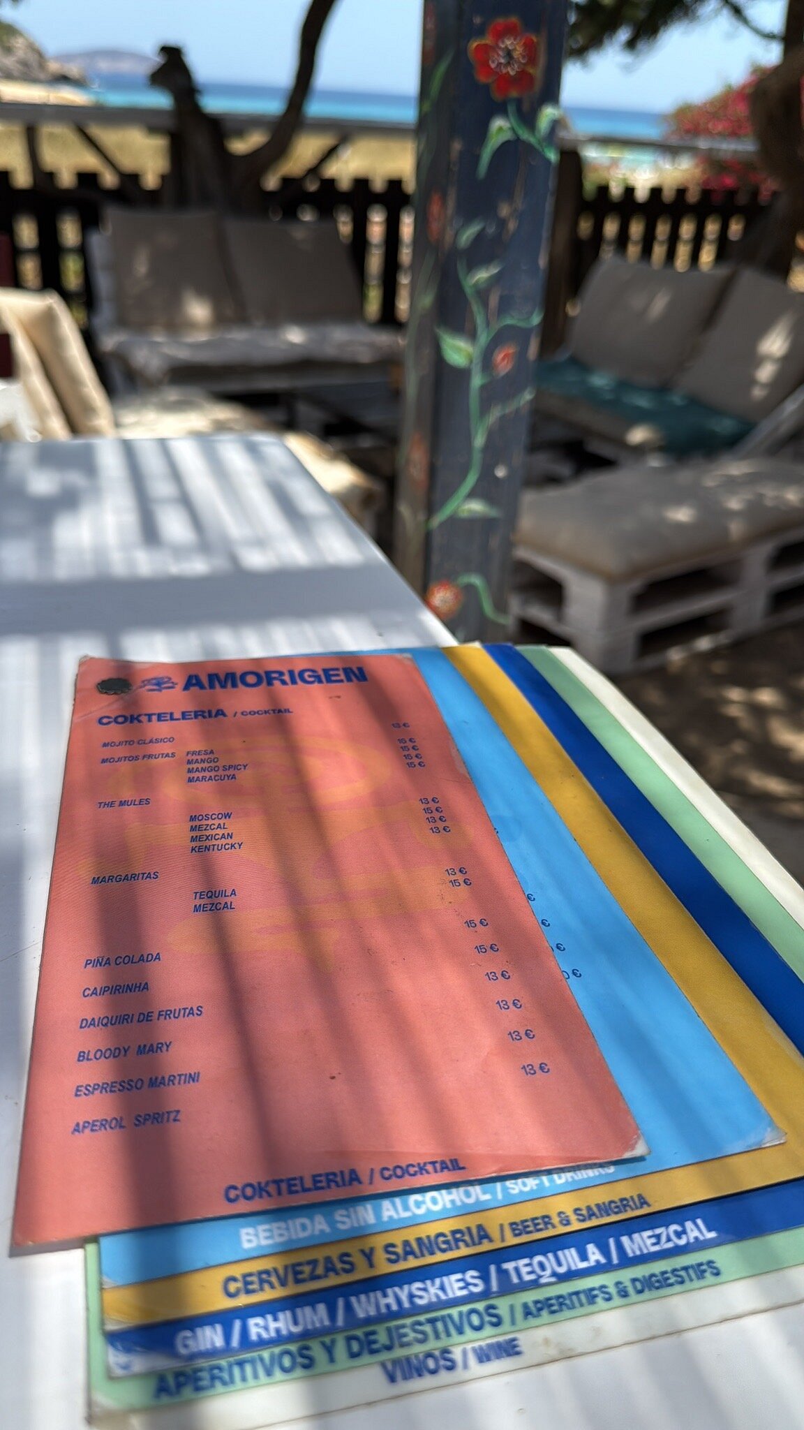 L'Amorigen Ibiza: het beschilderde strandrestaurant van Cala Nova 3 De menukaart van L'Amorigen op een witte tafel, met daarachter het houten terras met ligbedden en de begroeiing van de duinrand