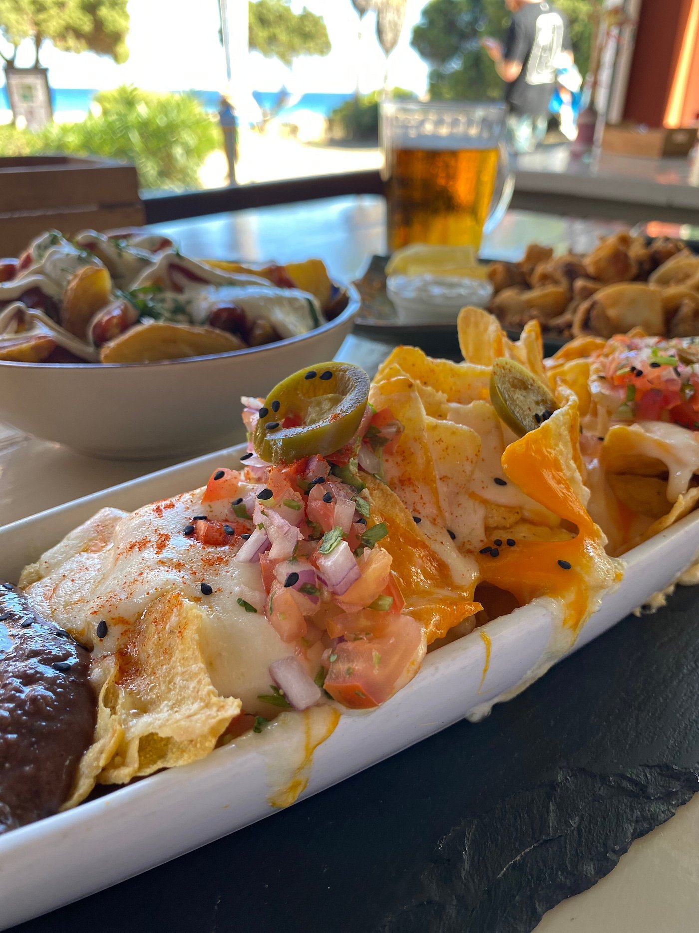 L'Amorigen Ibiza: het beschilderde strandrestaurant van Cala Nova 9 Een schaal nacho's met toppings en een glas bier op het terras van L'Amorigen, met palmen en de zee op de achtergrond