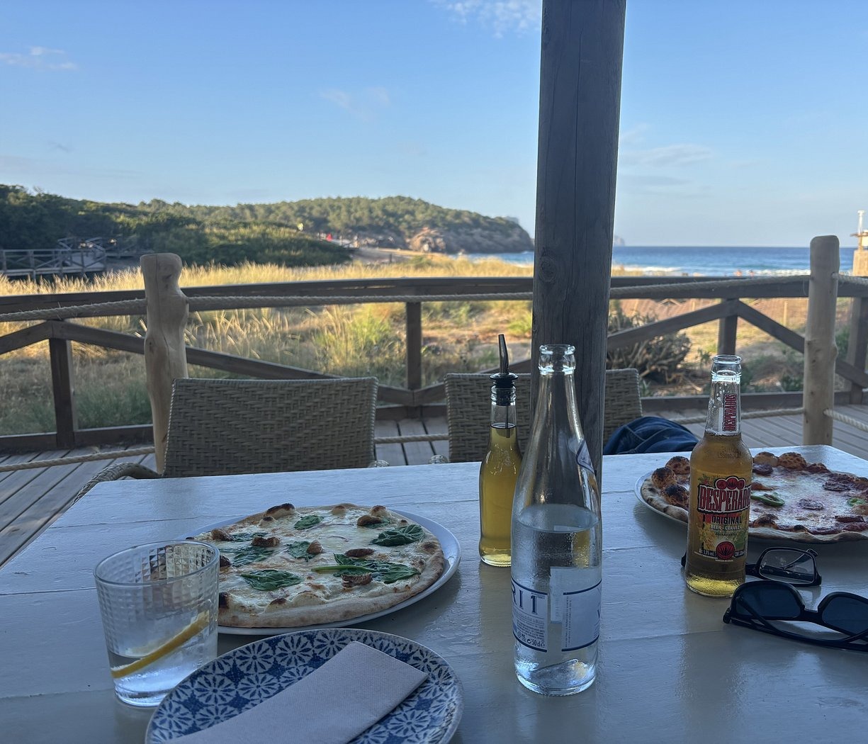 L'Amorigen Ibiza: het beschilderde strandrestaurant van Cala Nova 1 Twee pizza's op een witte tafel op het houten terras van L'Amorigen, met de rope-reling, het duingras en de baai van Cala Nova met de strandwachtpost erachter