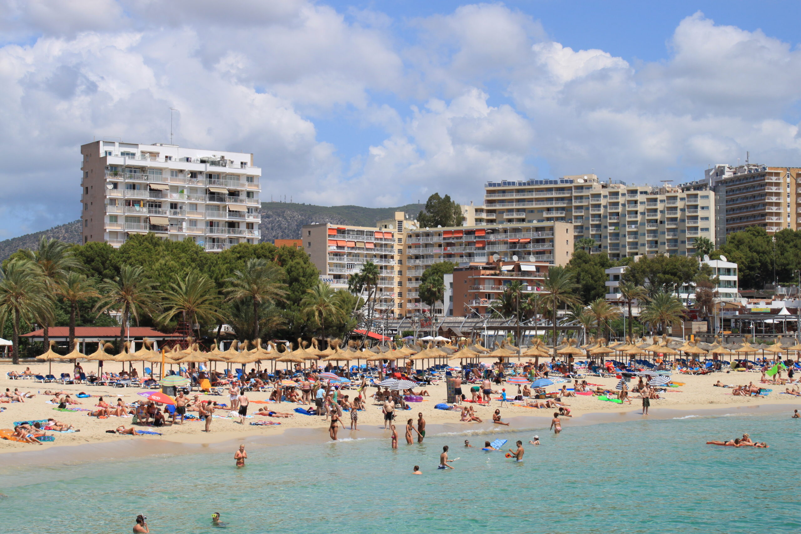 De Platja de Magaluf op een zomerdag vanaf het water gezien, met de rij rieten parasols, de palmen langs de promenade en de hotelblokken erachter