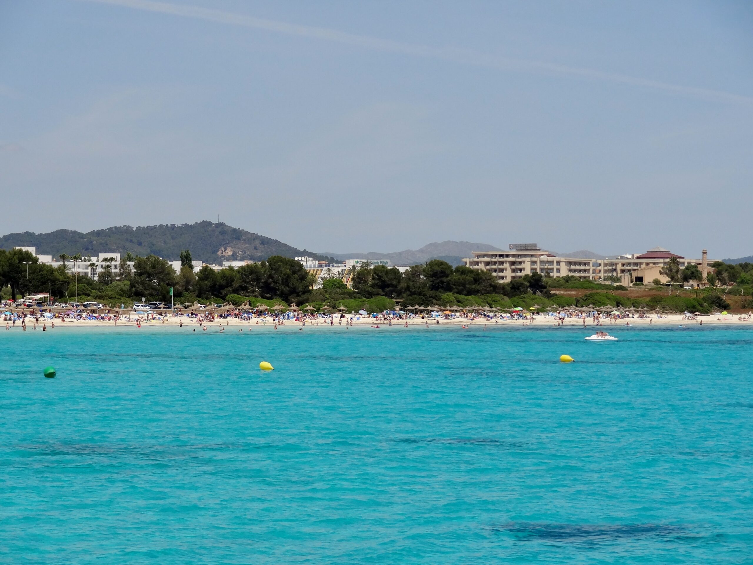 Het strand van Sa Coma gezien vanaf het water, met lichtturquoise ondiepten, gele zwemboeien en de beboste heuvels achter de badplaats