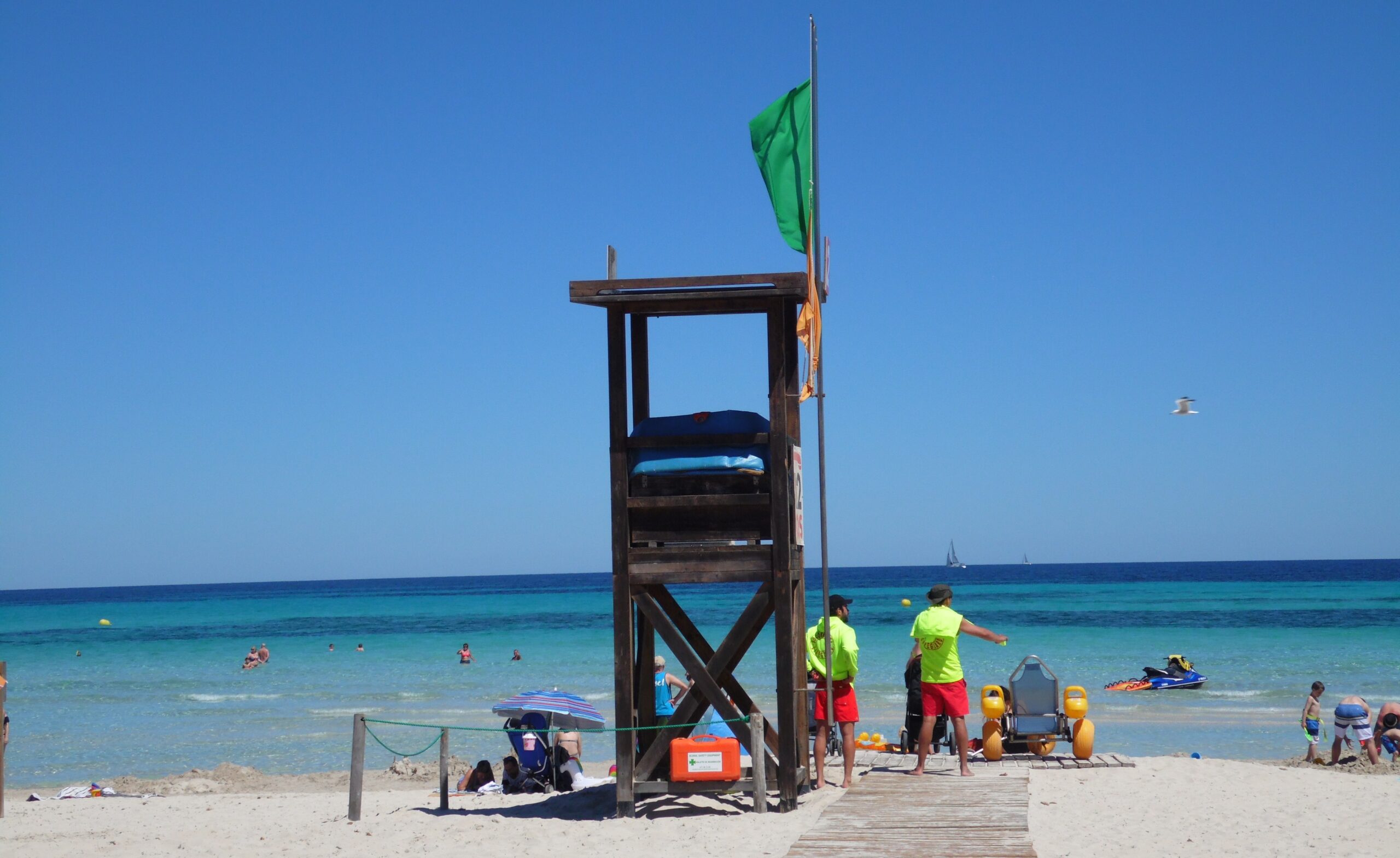 De houten reddingspost op het strand van Sa Coma met de groene vlag in top, een amfibische strandrolstoel ernaast en zwemmers in het ondiepe water