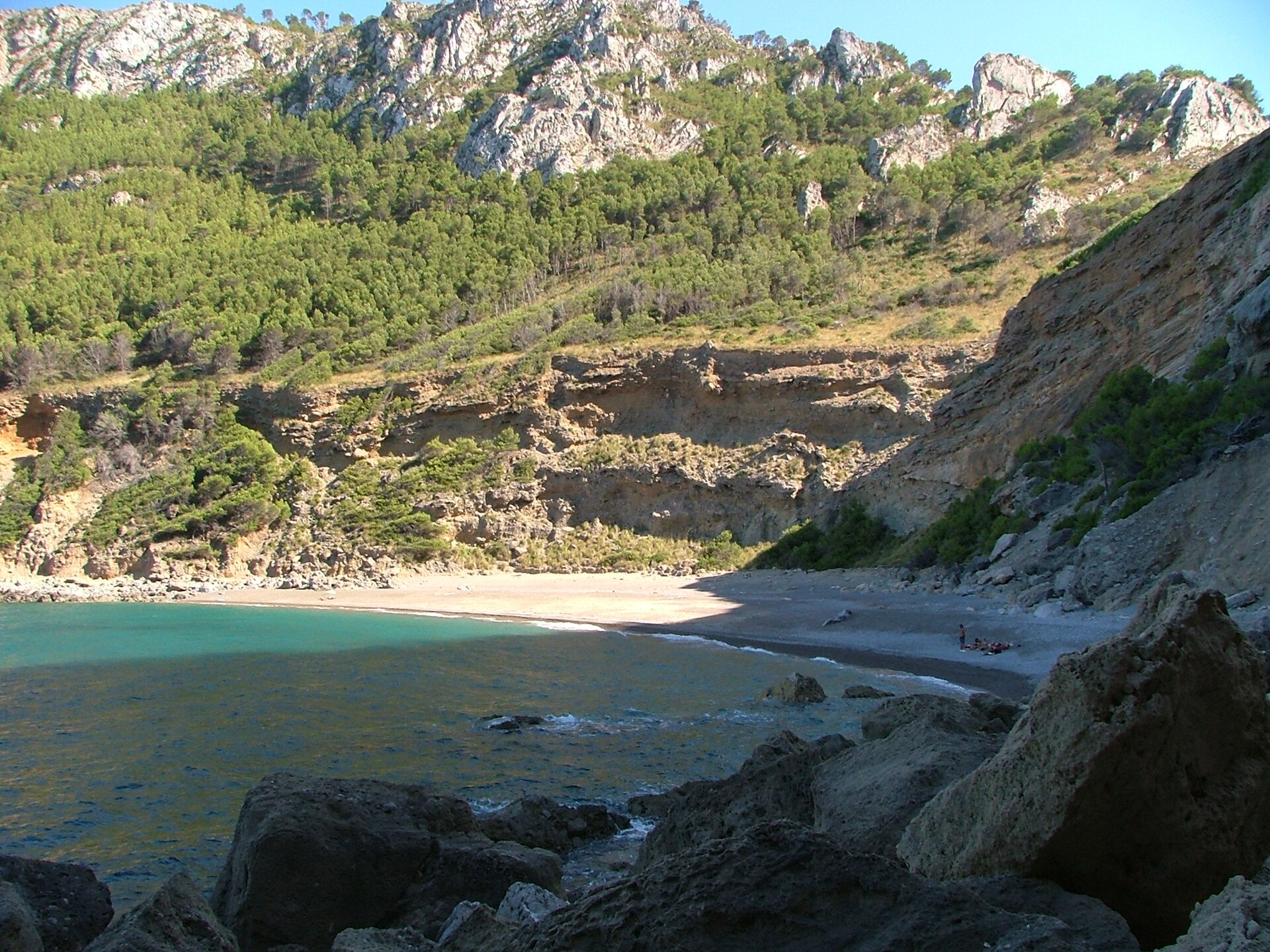 De baai van Coll Baix gezien vanaf de rotsen, met het beboste schiereiland erboven en enkele badgasten op het strand