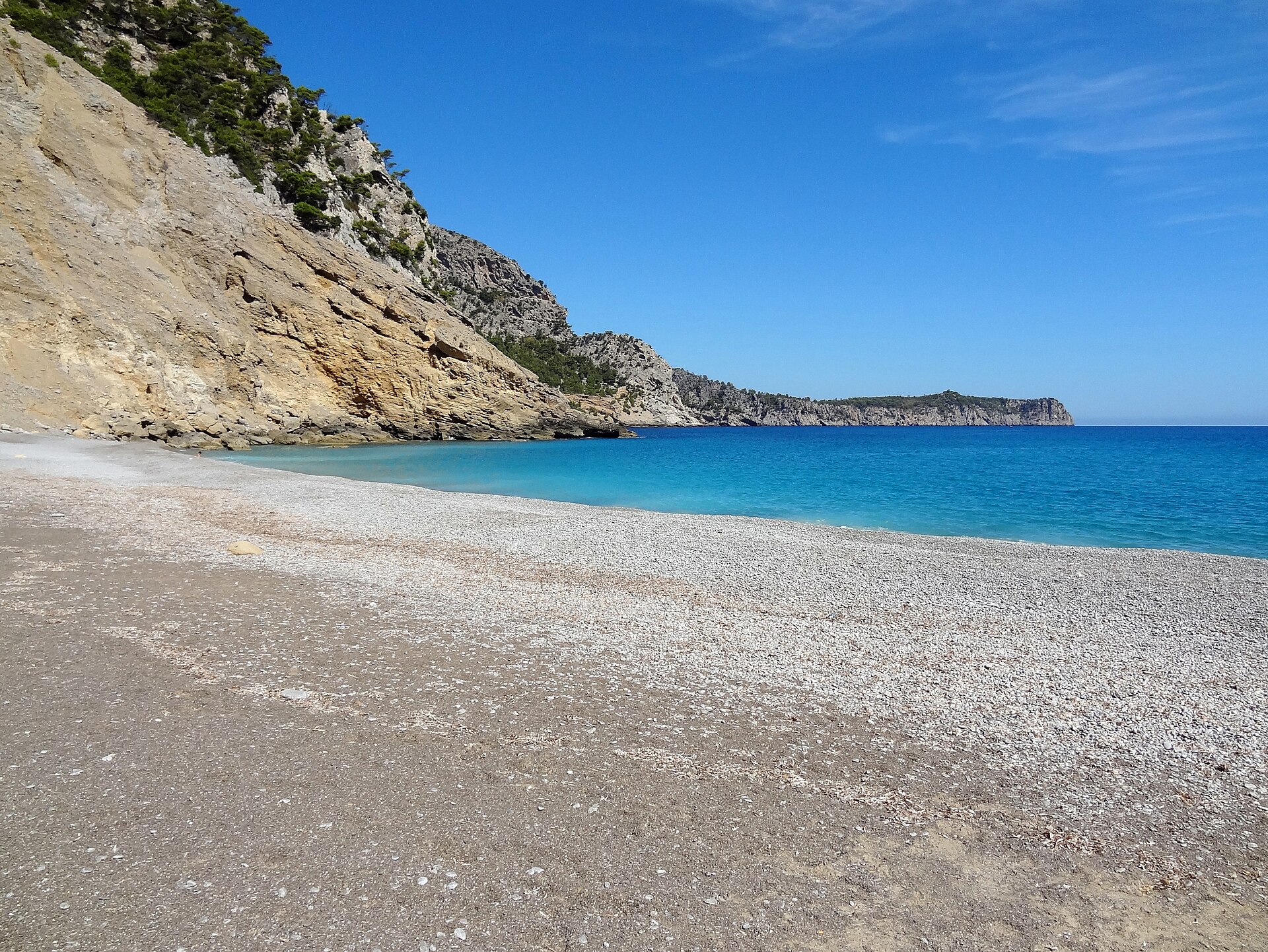 De lichte kiezels van Platja des Coll Baix van dichtbij, met het turquoise water en de kaap aan de horizon