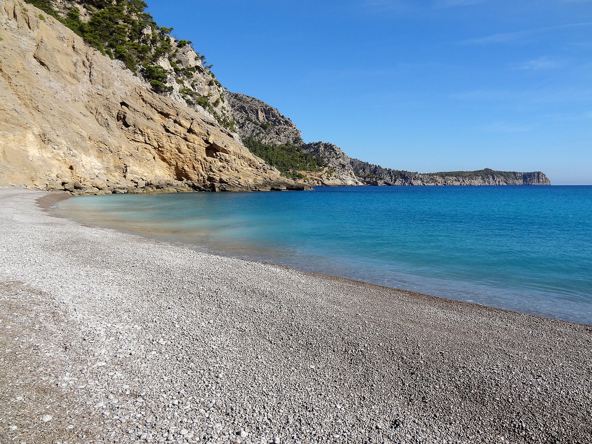 De boog van lichte kiezels van Platja des Coll Baix met turquoise water en de steile klif erachter