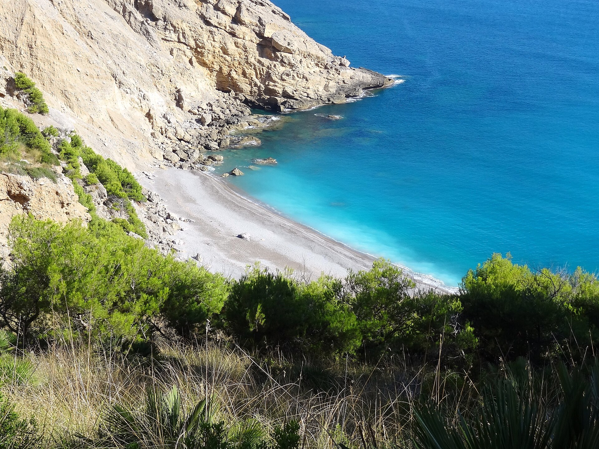 Platja des Coll Baix van bovenaf gezien, met de kiezelboog, het turquoise ondiepe water en de puinhelling onder de klif