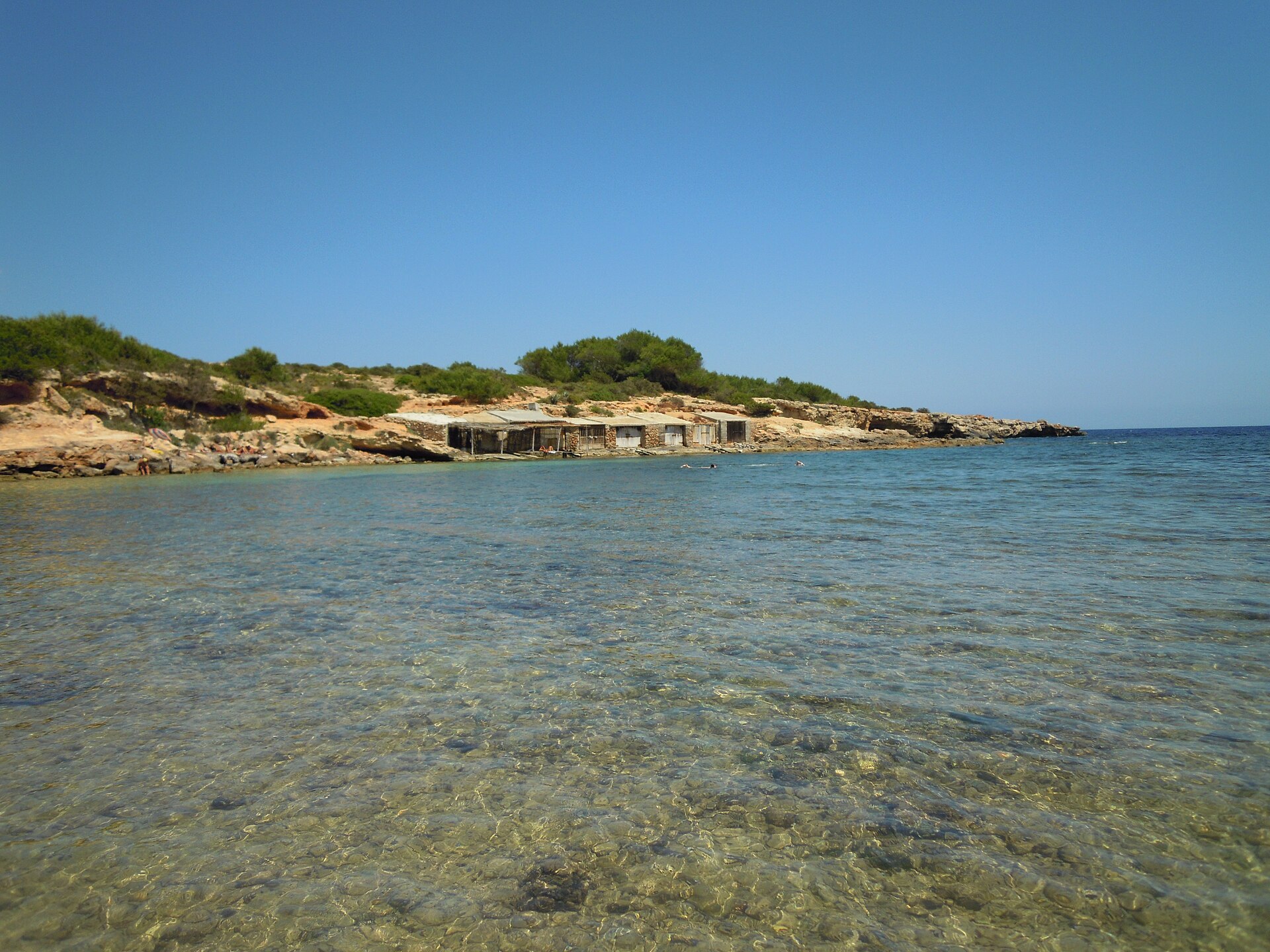 Platja de s’Estanyol Ibiza: de rotsbaai bij Cap Martinet 2 Rij oude stenen botenhuizen met houten deuren aan de overkant van de baai van s'Estanyol, met ondiep helder water ervoor