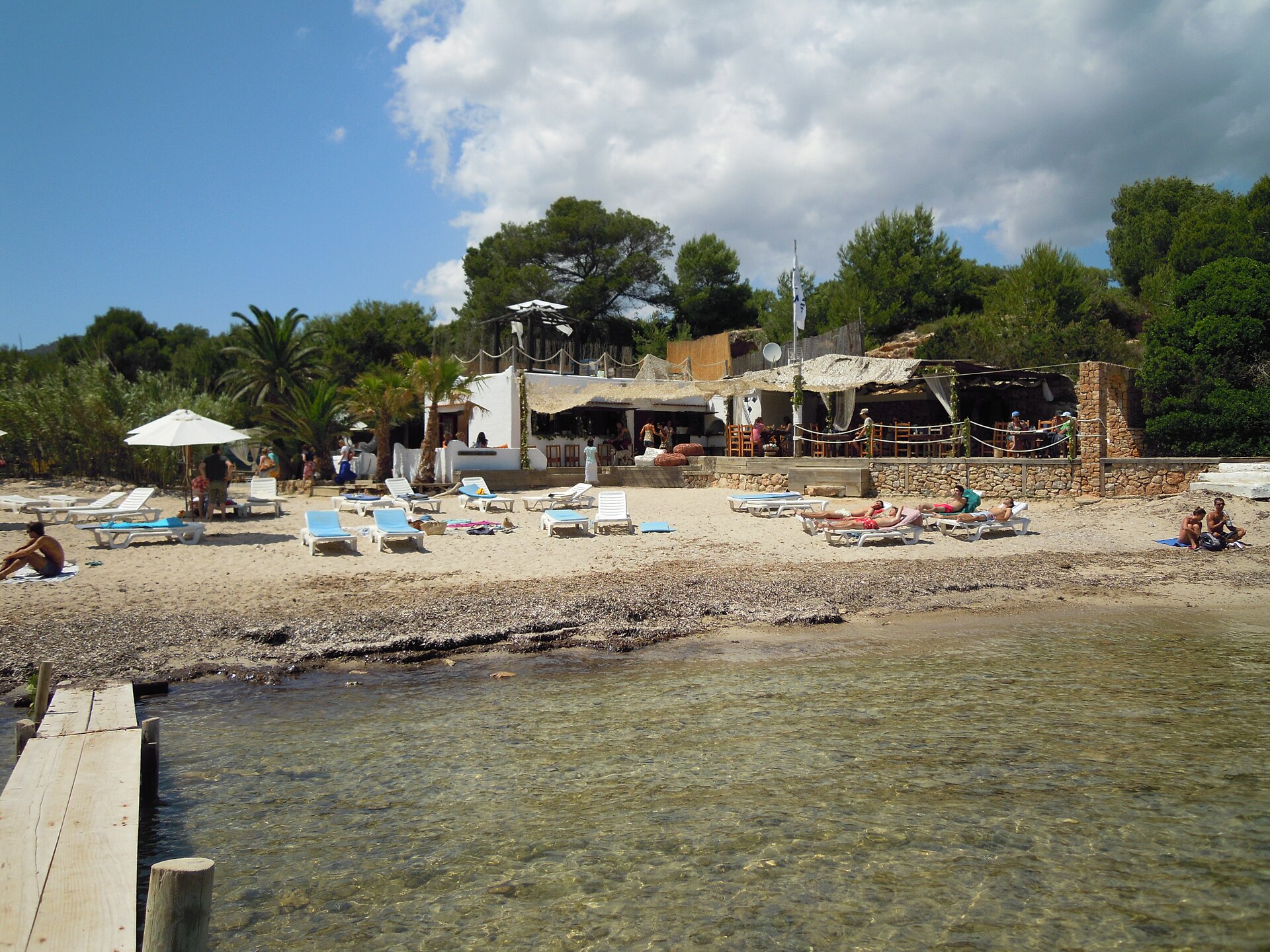 Platja de s’Estanyol Ibiza: de rotsbaai bij Cap Martinet 3 Het strandrestaurant aan s'Estanyol met terras, ligbedden op het zand en een houten steiger op de voorgrond