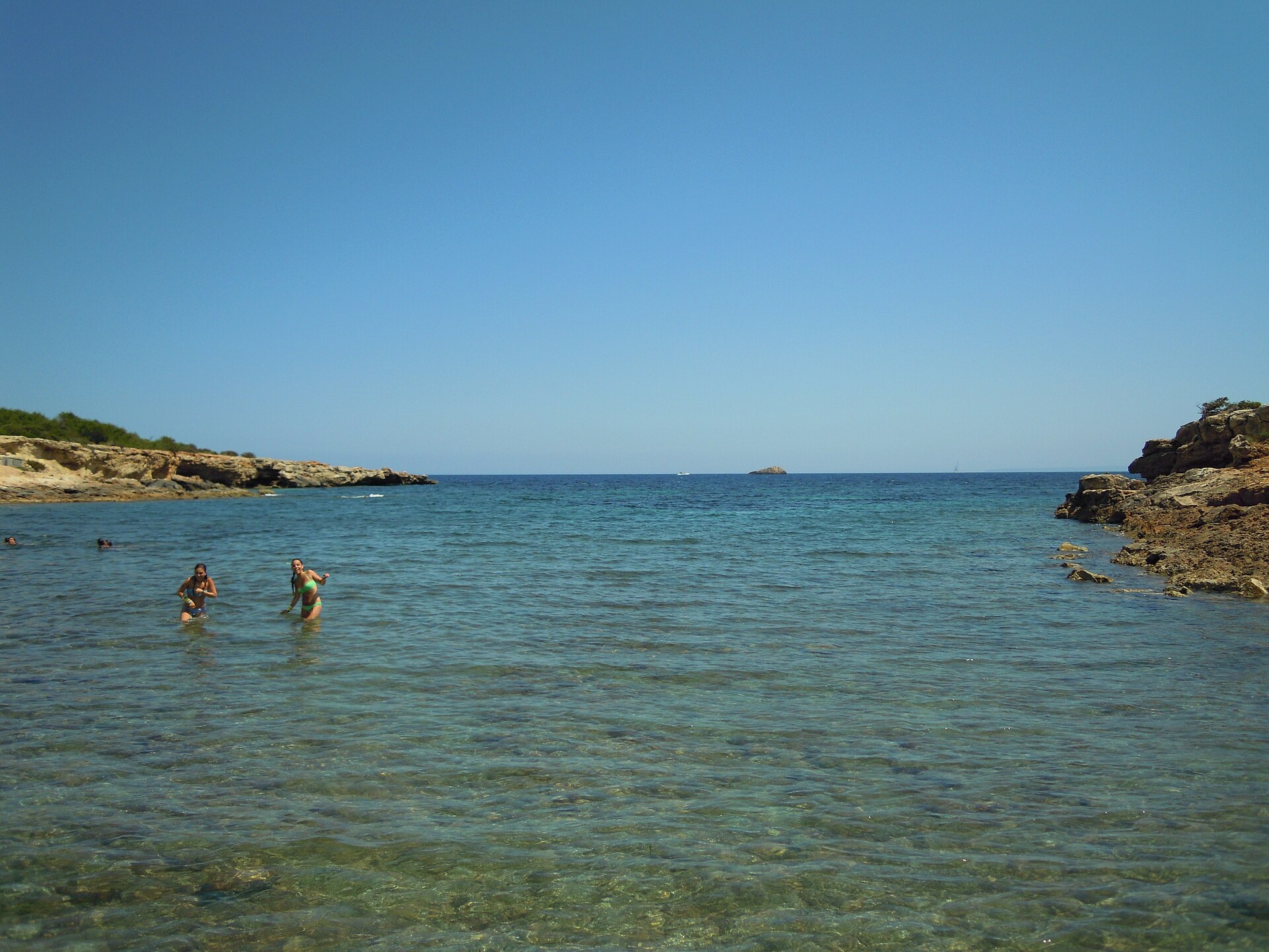 Platja de s’Estanyol Ibiza: de rotsbaai bij Cap Martinet 5 Zwemmers in het ondiepe heldere water van s'Estanyol met de rotsige baaimond en een eilandje aan de horizon