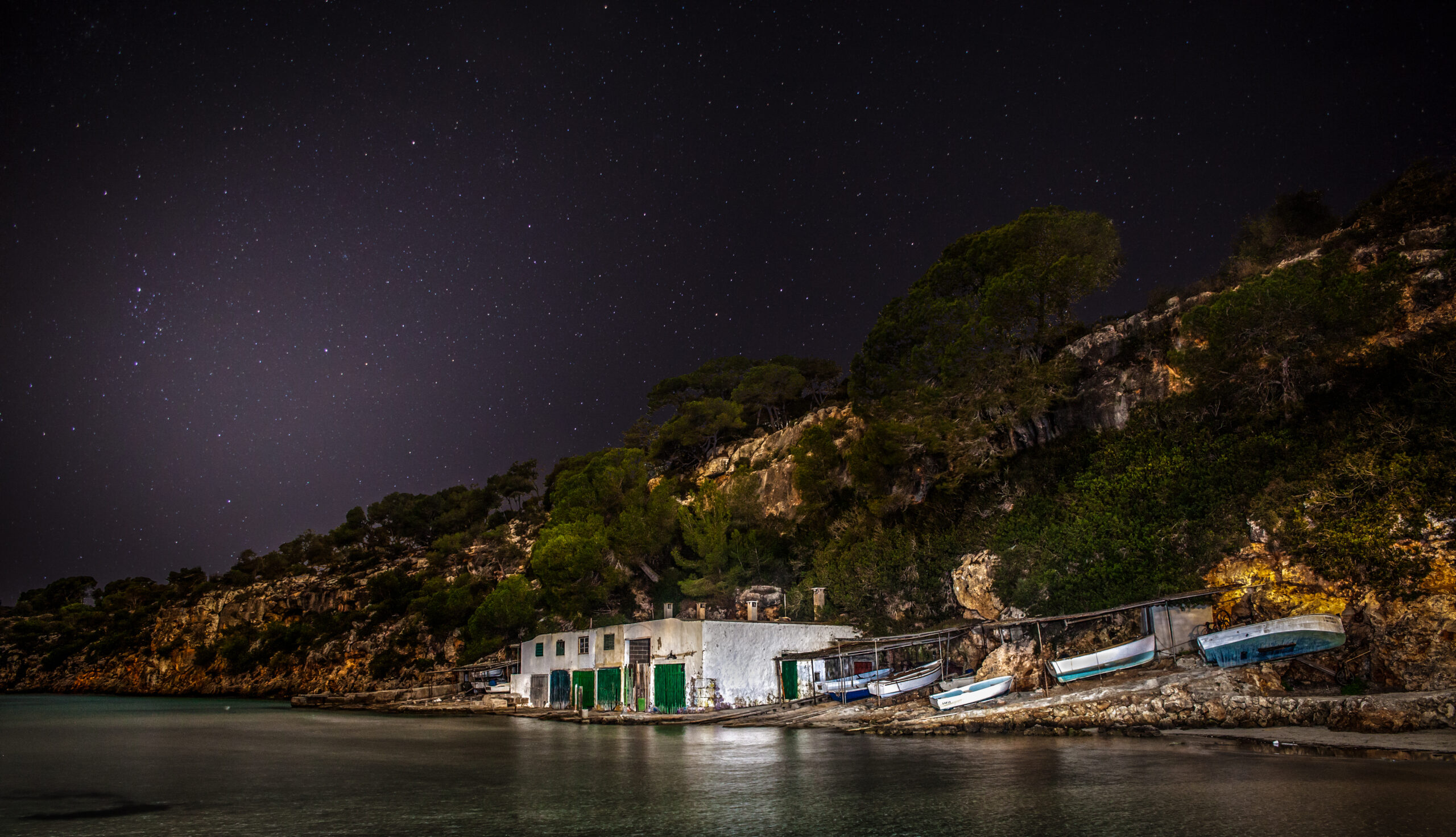 De boothuizen van Cala Pi bij nacht onder een sterrenhemel, met opgetrokken bootjes op de hellingen en de beboste kliffen erachter